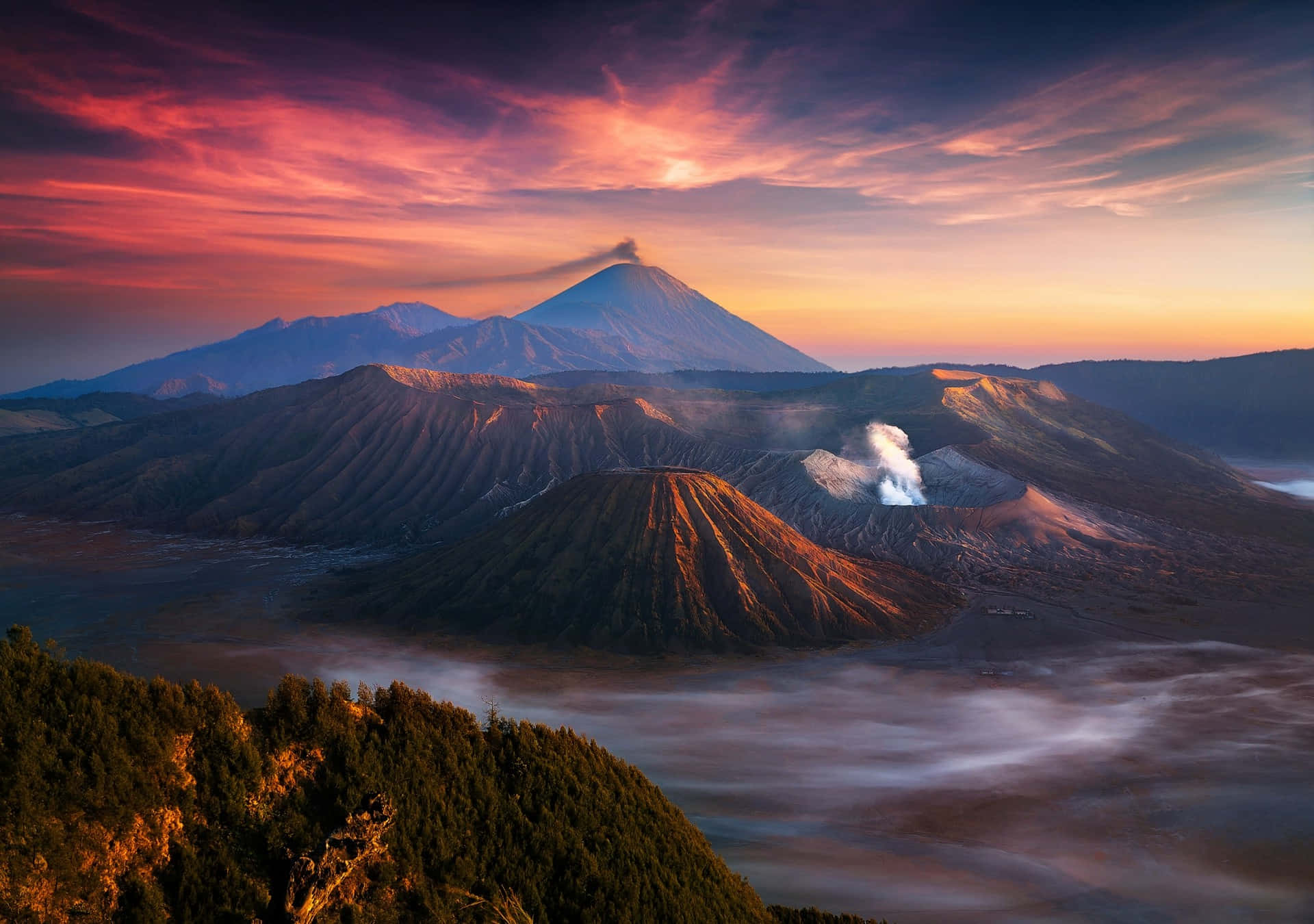 Indonesia 桌布 壁紙 — 日出時分的雄偉火山景觀，霧氣繚繞的山脈和冒著蒸汽的火山口上方，是充滿活力的粉紅色和橙色天空。