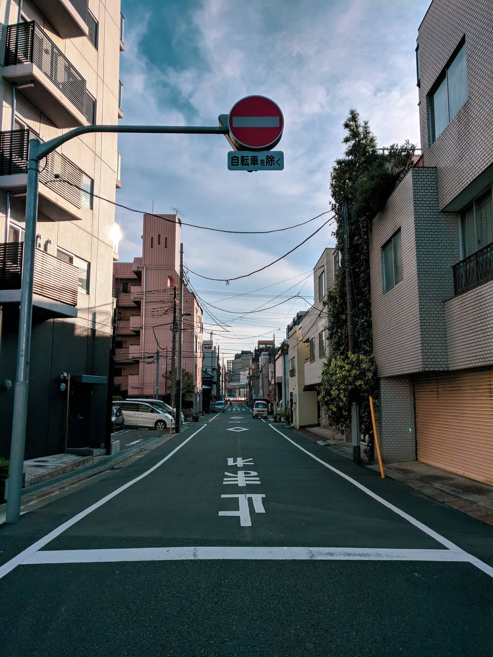 一條狹窄的日本街道，有建築物、停放的汽車和藍天下的一個禁止進入標誌。