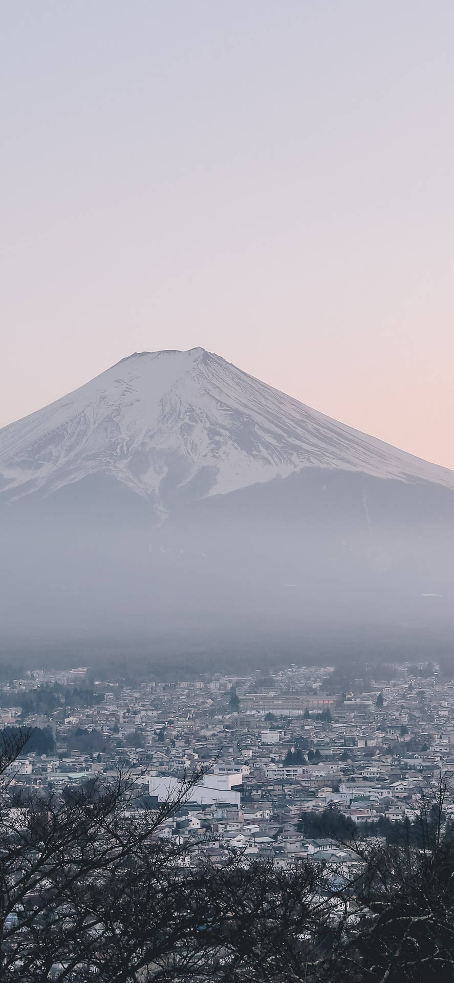 日式美學 iPhone 著名富士山 壁紙 — 白雪覆蓋的富士山在黎明時分，以柔和的粉紅色和灰色調，雄偉地聳立在霧氣瀰漫的小鎮之上。