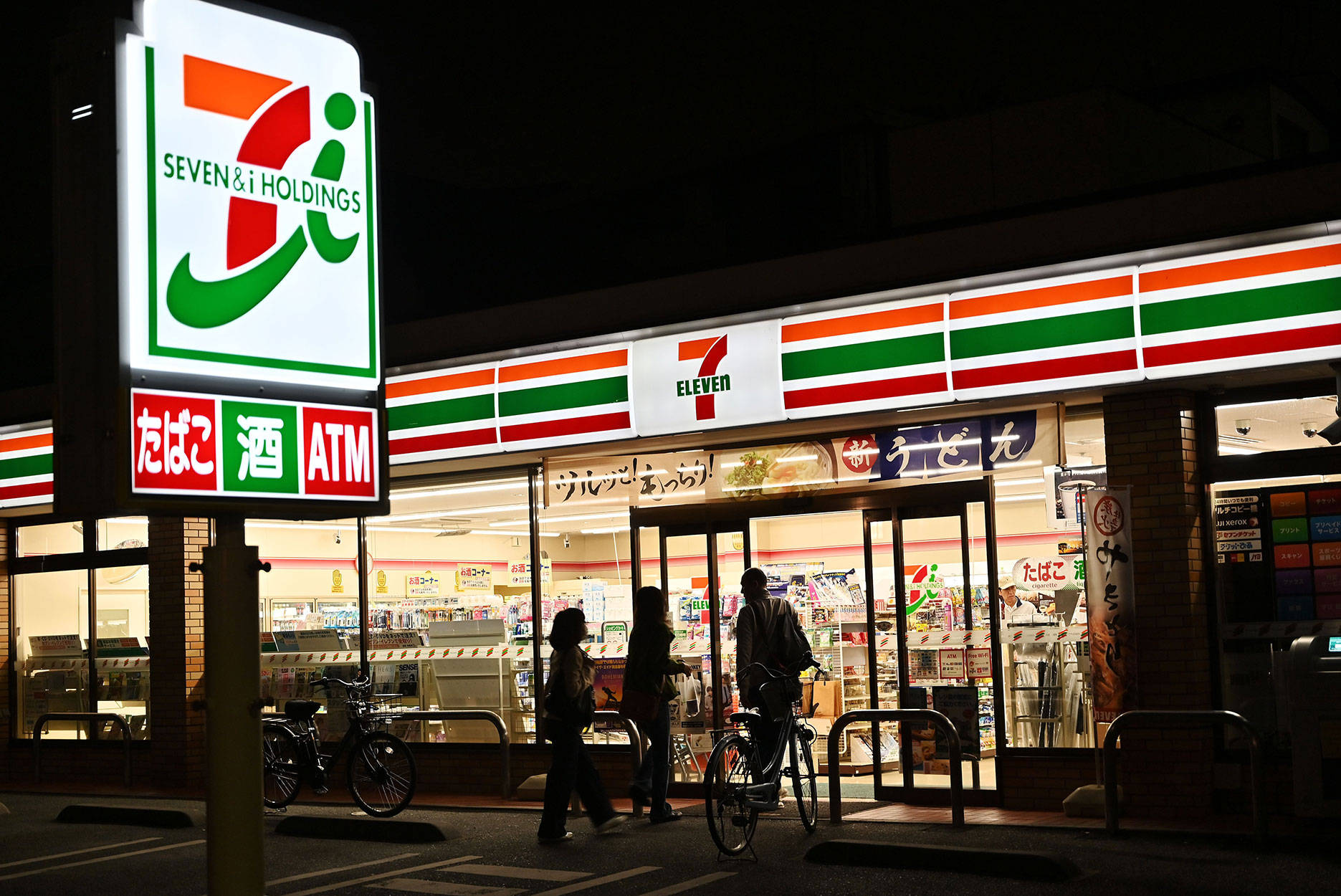 日本7-Eleven 壁紙 — 夜晚場景，一間燈火通明的7 Eleven便利店，有行人和單車經過。