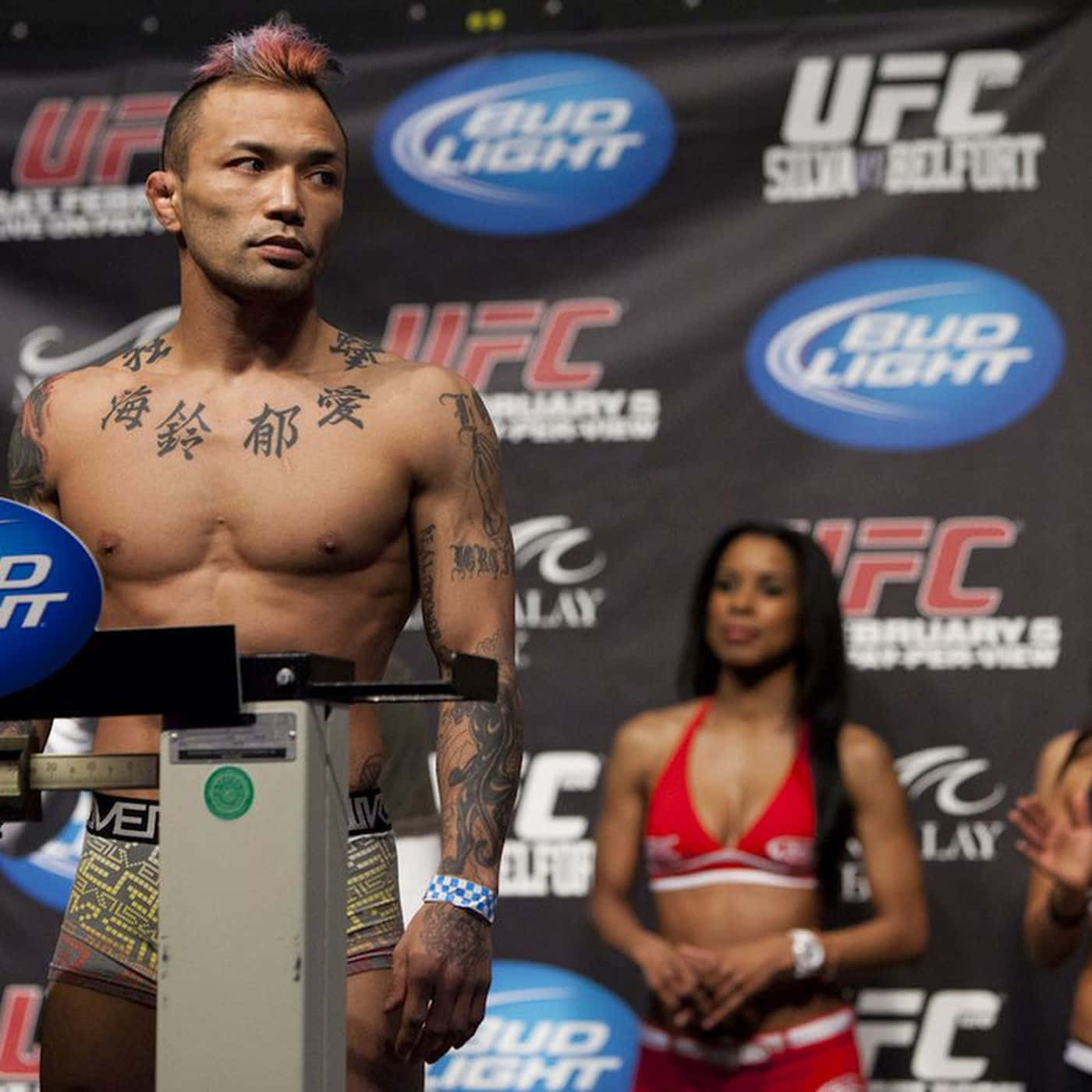 日本MMA傳奇Norifumi Yamamoto UFC 126體重秤亮相 壁紙 — 一位身上有紋身、留著粉紅色莫霍克頭的UFC拳手在稱重活動中站在體重秤上，背景中有一位身穿比基尼的擂台女郎。