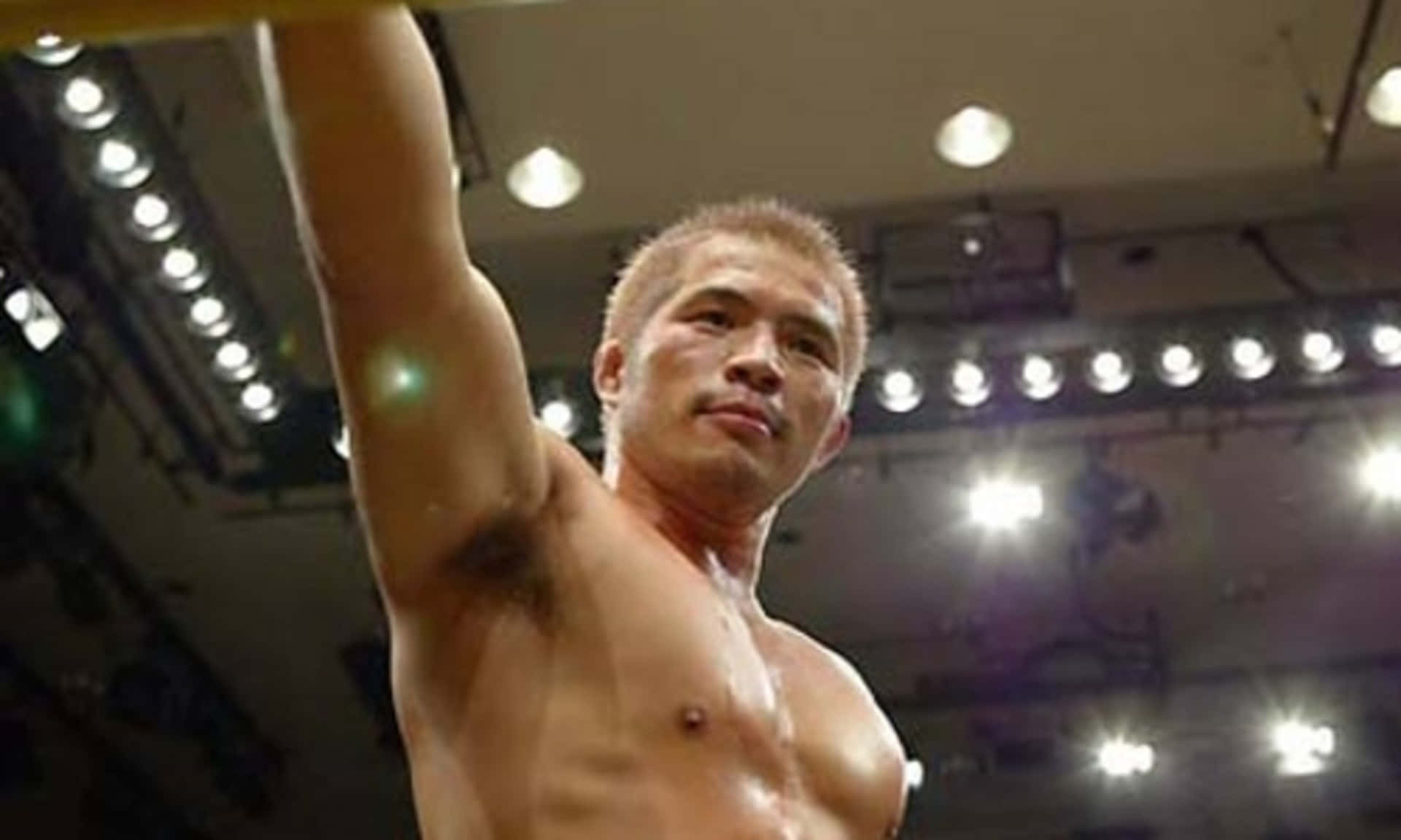 日本MMA選手Hayato Sakurai低角度拍攝 壁紙 — 一名赤裸上身的肌肉戰士在明亮的競技場燈光下勝利地舉起手臂。