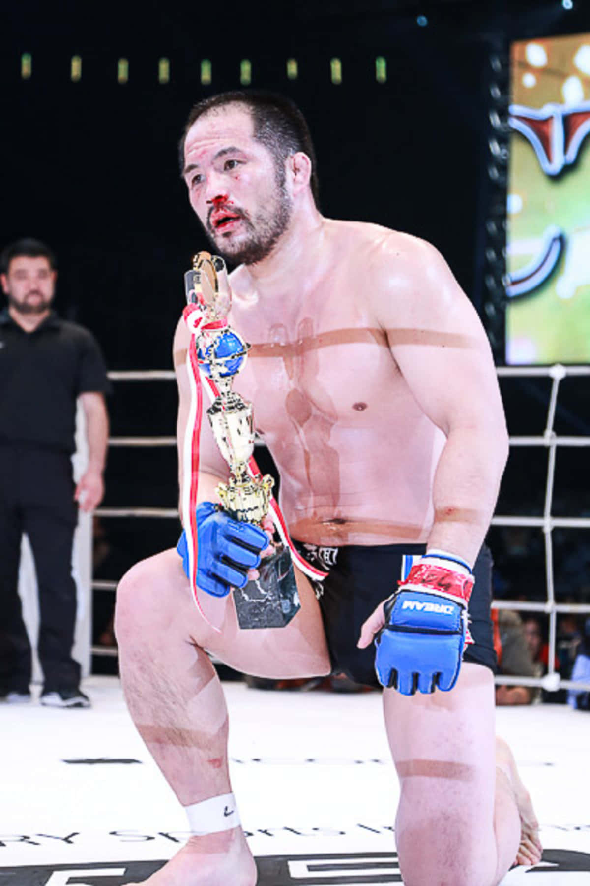 日本MMA選手Hayato Sakurai血跡滿面 壁紙 — 一名血跡斑斑的MMA拳手在八角籠中跪地，手持冠軍獎盃，戴著藍色手套，軀幹上纏著膠帶。