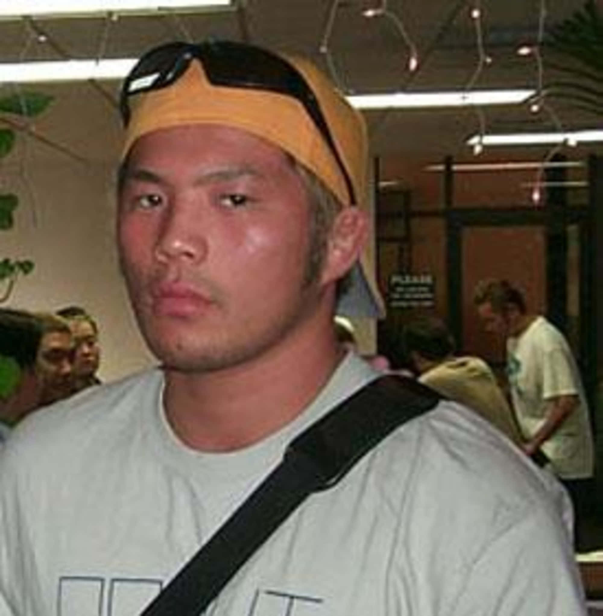 日本MMA選手Hayato Sakurai中角度拍攝 壁紙 — 一個戴著黃色鴨舌帽和太陽眼鏡擱在頭上的男人，在光線明亮的室內環境中直視鏡頭。