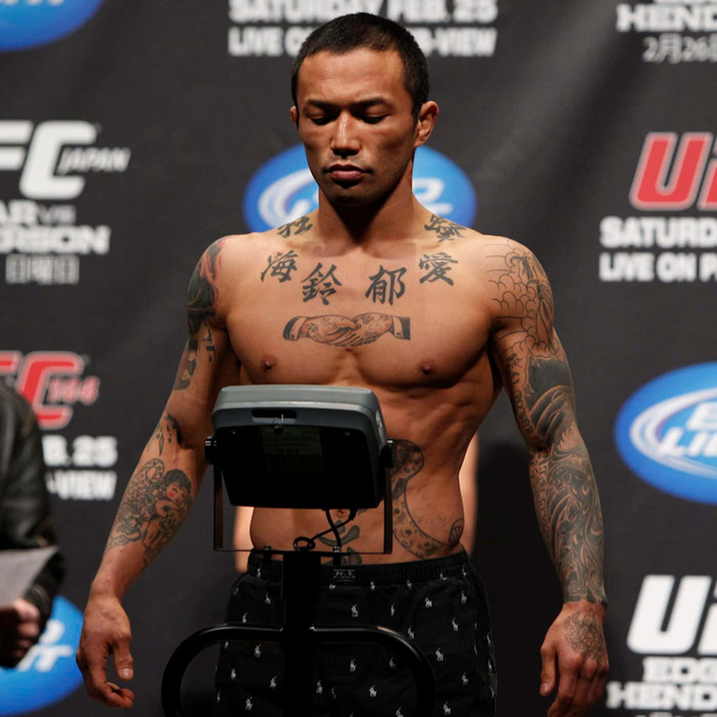 日本UFC選手Norifumi Yamamoto UFC 144體重秤亮相 壁紙 — 一位有紋身的UFC拳手在品牌背景的稱重活動中赤膊站在體重秤上。