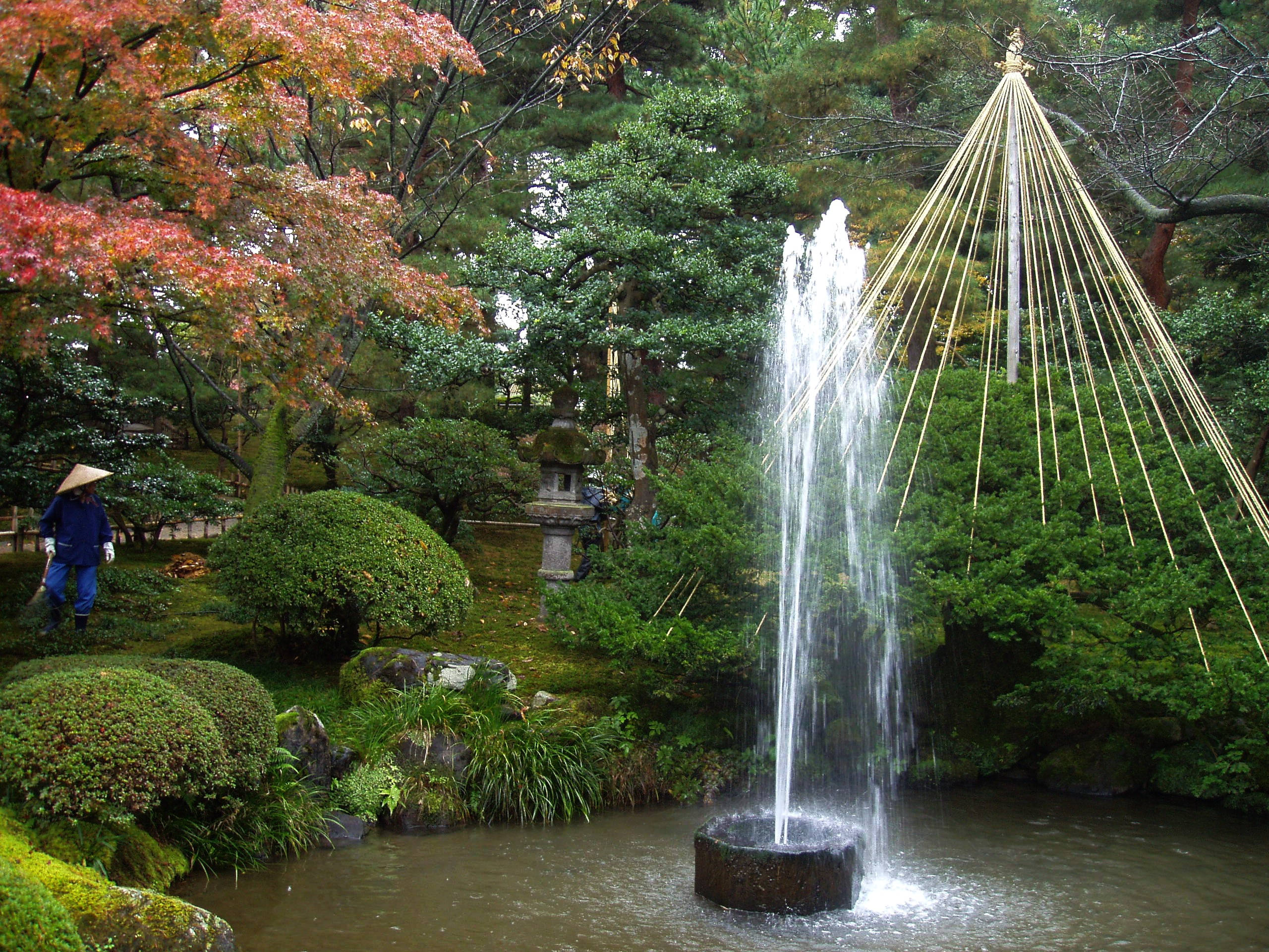 日本兼六園 壁紙 — 一座寧靜的日本花園噴泉傾瀉入池塘，周圍環繞著秋葉和長滿苔蘚的岩石。