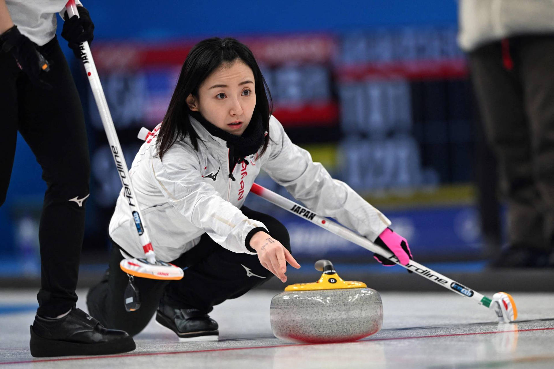 日本冰壺隊成員 Fujisawa Satsuki 壁紙 — 一位專注的日本冰壺運動員身穿白色外套，以高度集中力將花崗岩冰壺石推過冰面。