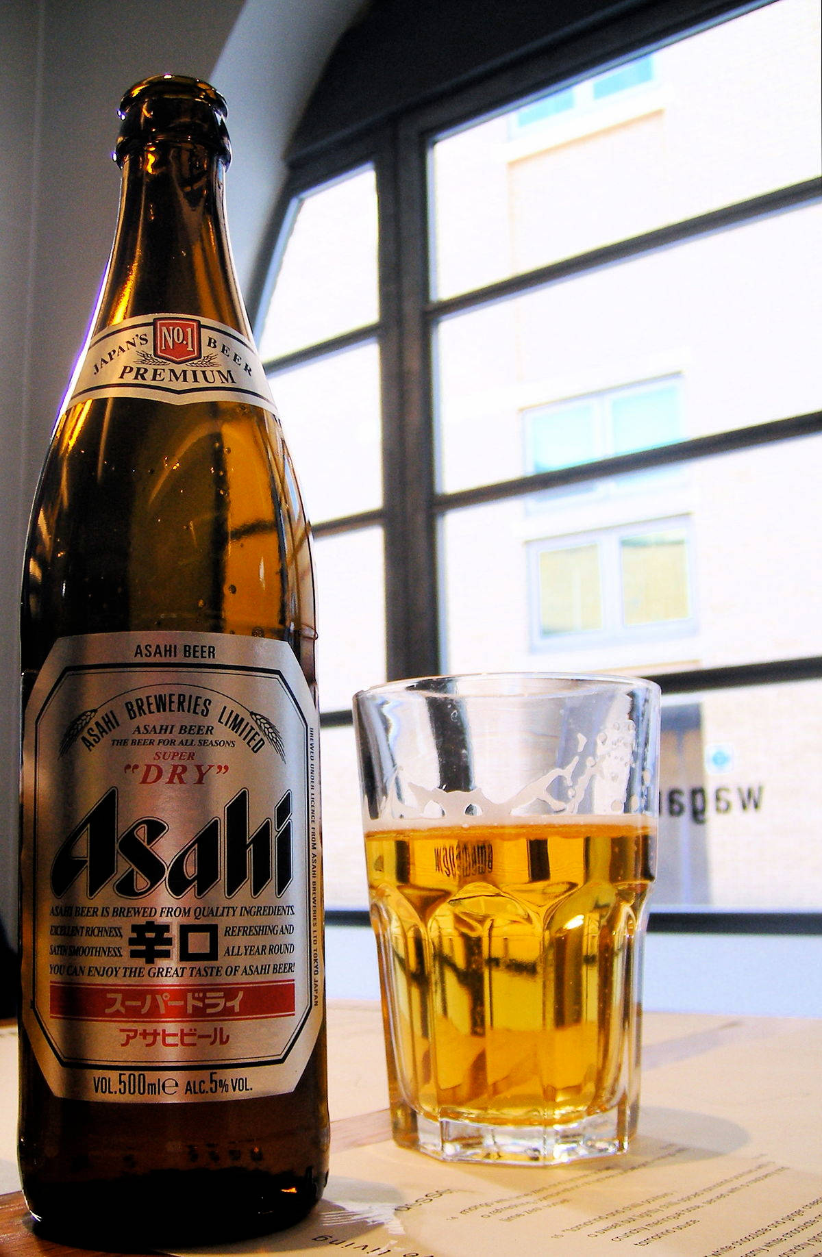 日本啤酒廠朝日超乾 壁紙 — 一支冰凍的朝日超乾啤酒和一個滿杯放在窗邊的桌子上，窗外可見城市建築。