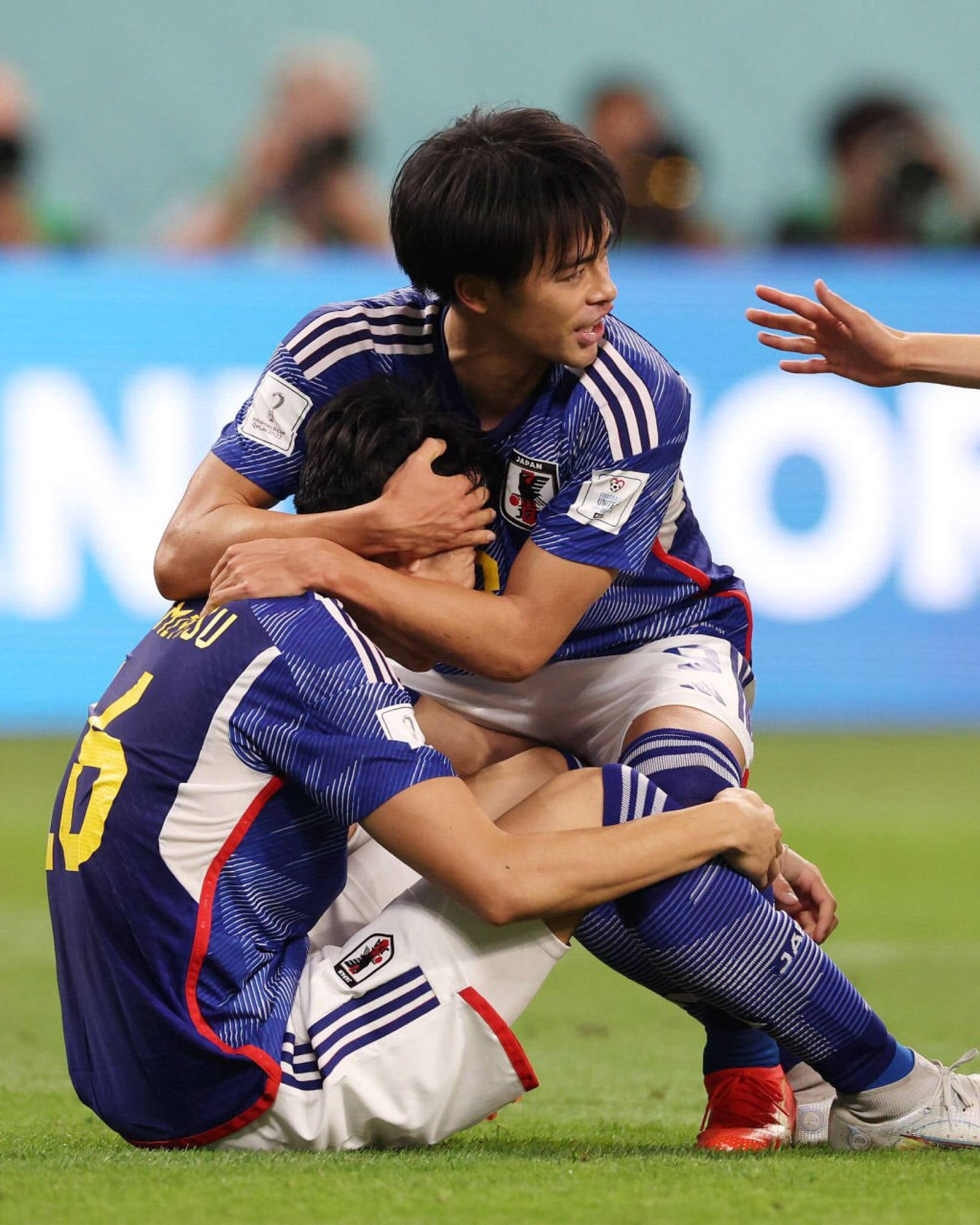 日本國家足球隊Mitoma安慰Tomiyasu 壁紙 — 兩名日本足球運動員在體育場燈光下，於充滿活力的綠色球場上情感相擁。