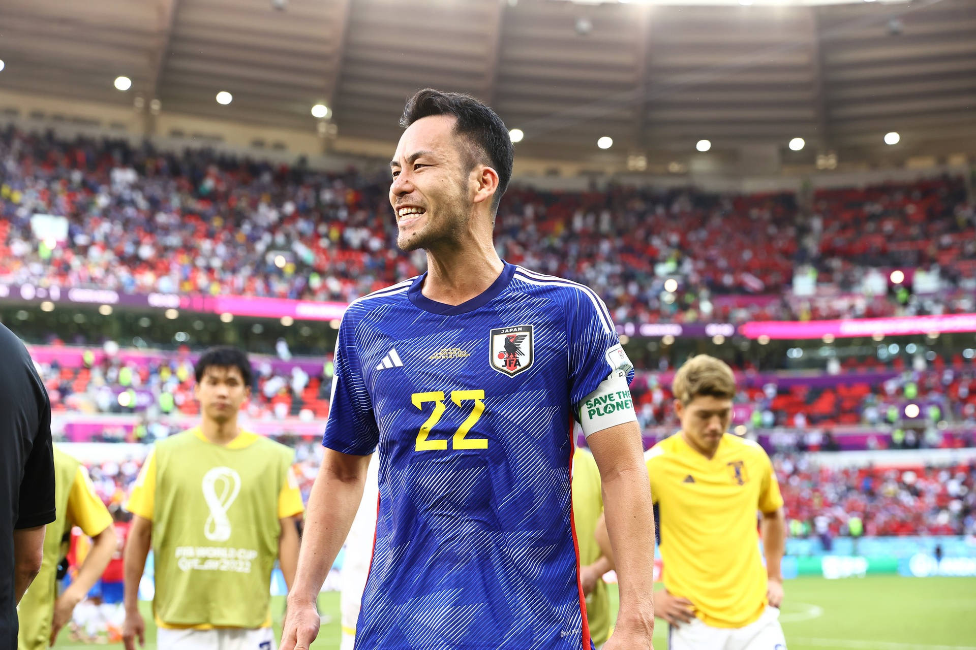 日本國家足球隊隊員吉田麻也賽後開心笑容 壁紙 — 一位身穿藍色22號球衣的日本足球員在球場上燦爛微笑，背景是歡呼的球迷。
