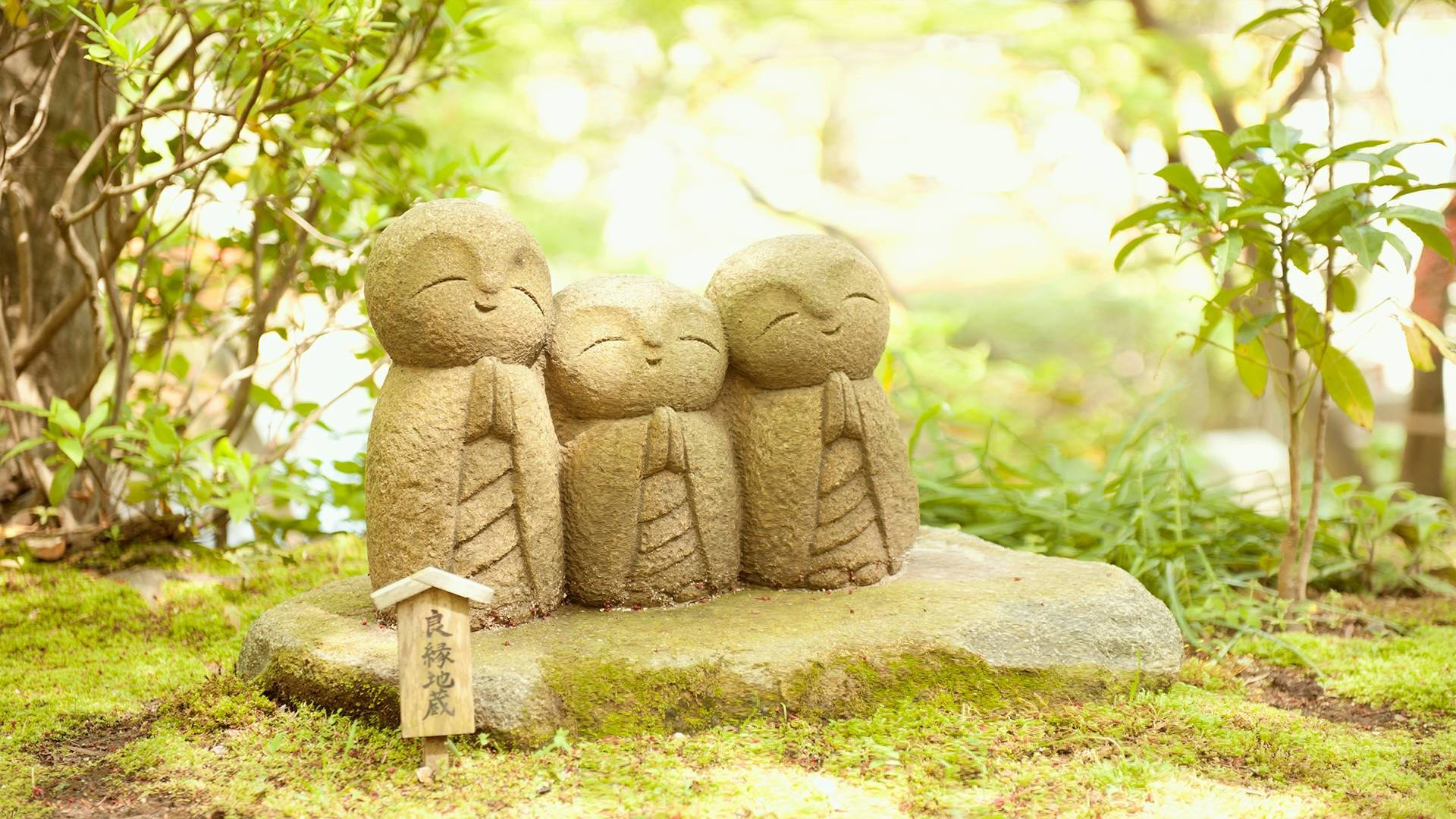 日本地藏菩薩雕像佛像 HD 壁紙 — 三個微笑的小孩石像並排站在陽光普照的花園中一塊長滿苔蘚的石頭上，四周綠意盎然。