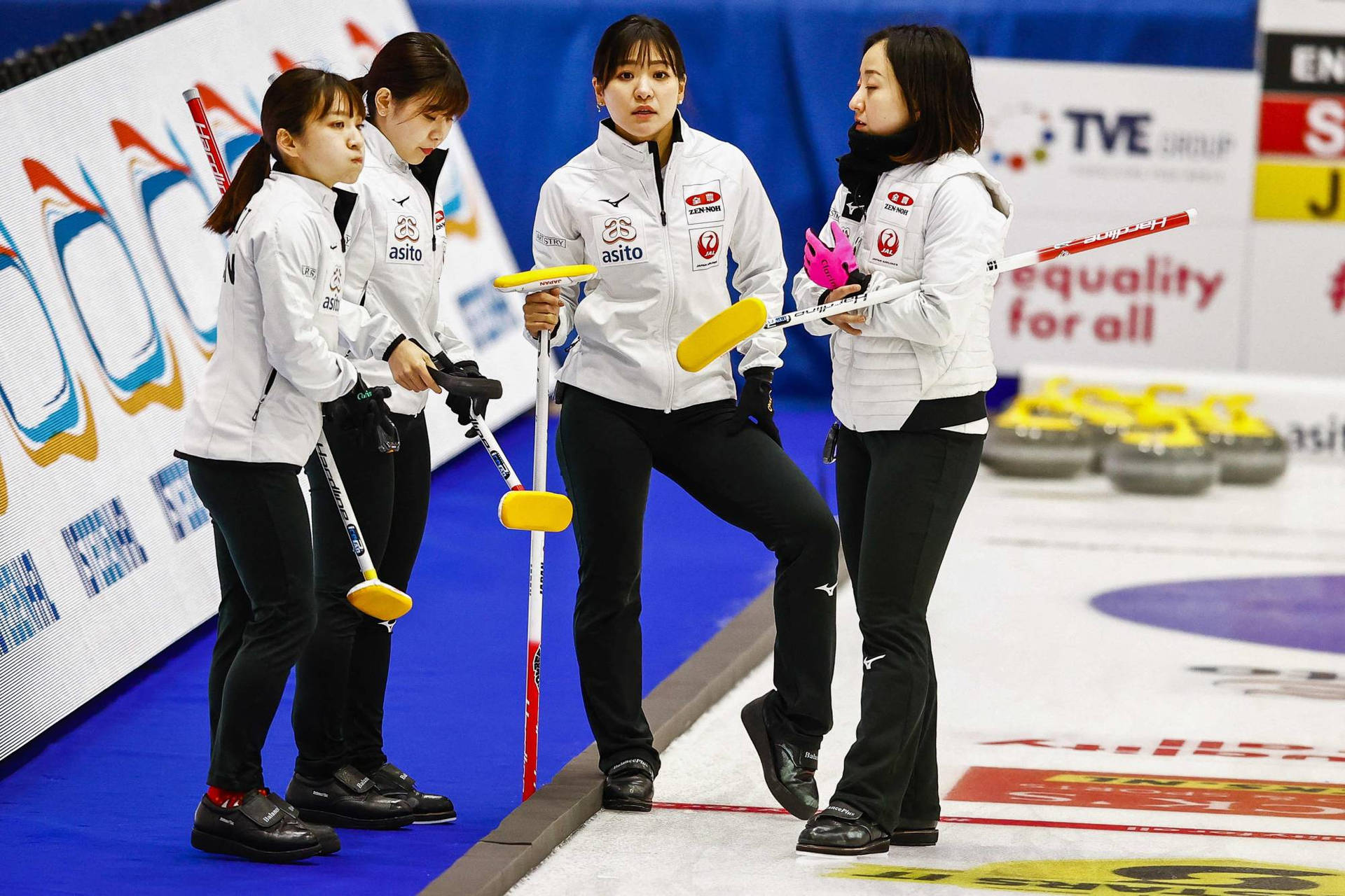 日本女子冰壺隊 壁紙 — 四位身穿白色制服的女冰壺運動員在冰場上拿著黃色冰壺掃帚激烈地制定策略。
