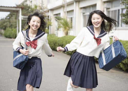 兩名身穿水手服、背著背包的女學生笑著在校園小徑上奔跑。