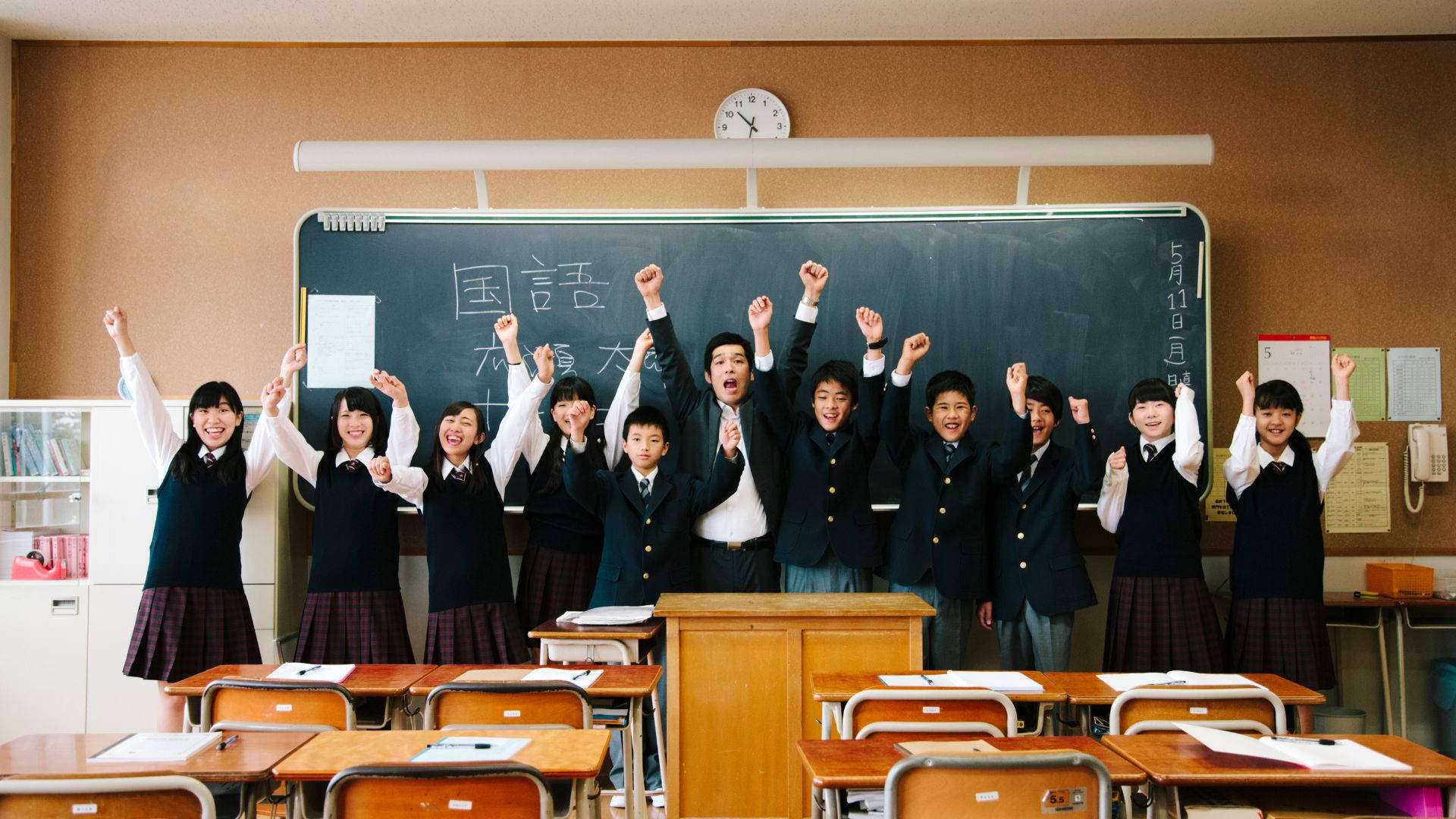 日本學校教室學童合影 壁紙 — 一群身穿制服、歡樂的日本學童在明亮的教室裡勝利地舉起拳頭。