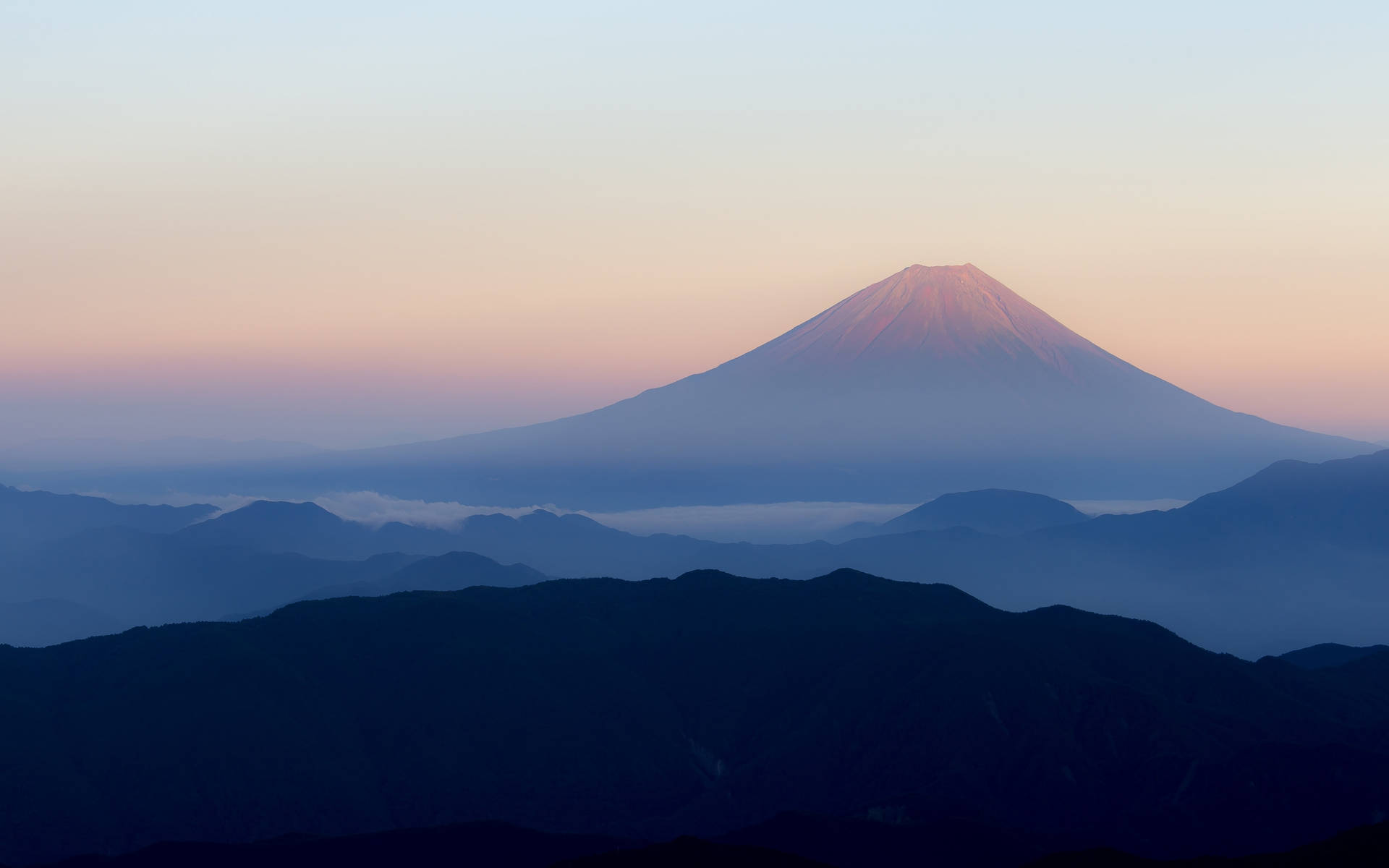 日本富士山日出 4K 壁紙 — 富士山在黎明時分雄偉地升起，柔和的粉紅色和藍色色調在層疊的山巒輪廓上空繪出天空。