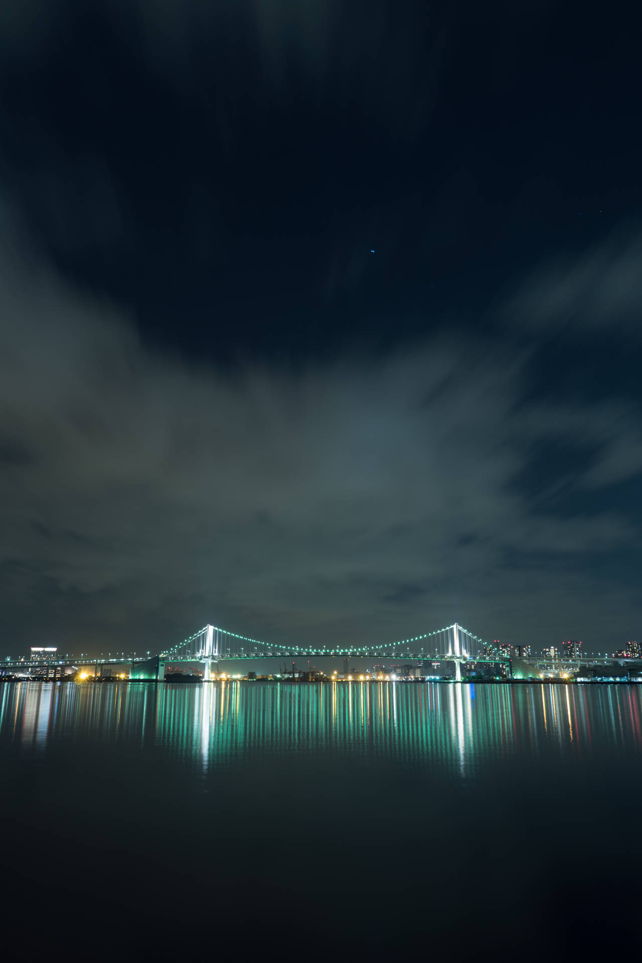 夜晚一座燈火通明的橋樑，其倒影映在雲天下的水面上。