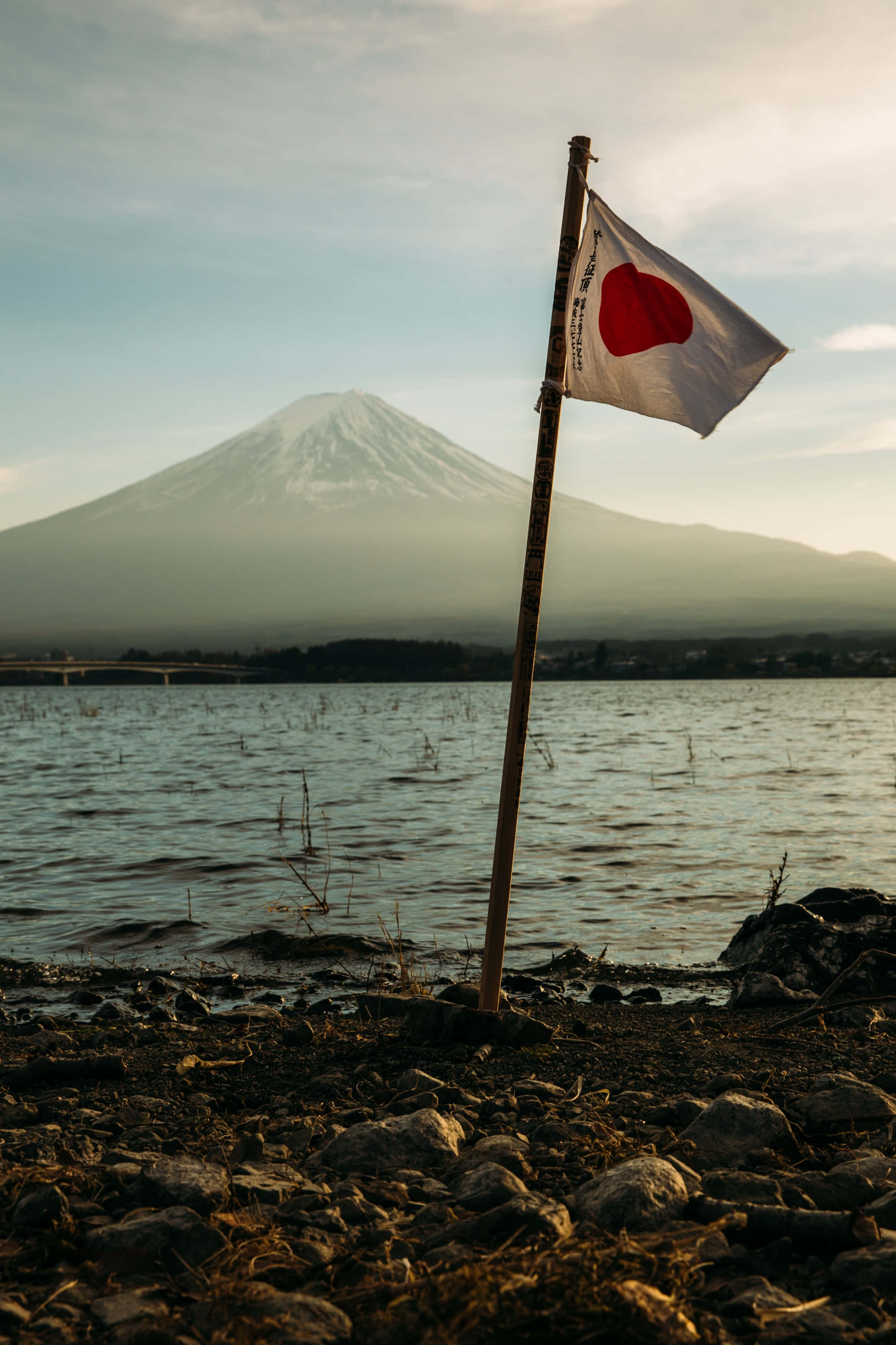 日本白雅致：白色之路的寧靜之美 壁紙 — 一支日本國旗在湖岸的棍子上飄揚，背景是夕陽下積雪的富士山。