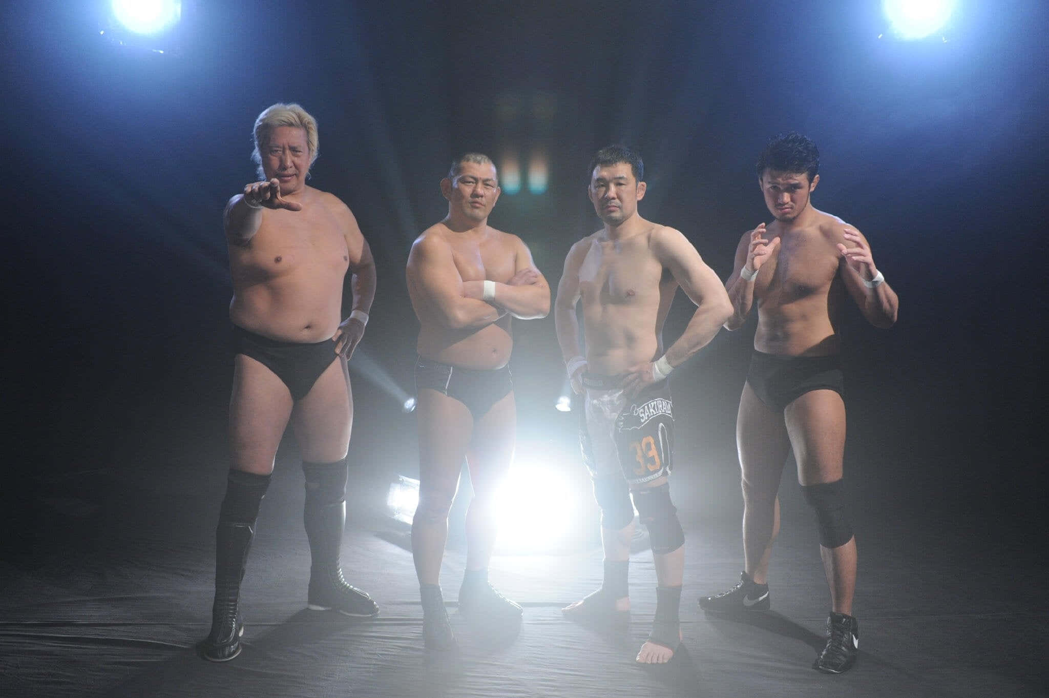 日本知名MMA選手櫻庭和志與其他日本職業摔角手合照 壁紙 — 四位肌肉發達的摔角手穿著黑色緊身褲，在黑暗的擂台上自信地擺姿勢，舞台燈光明亮。