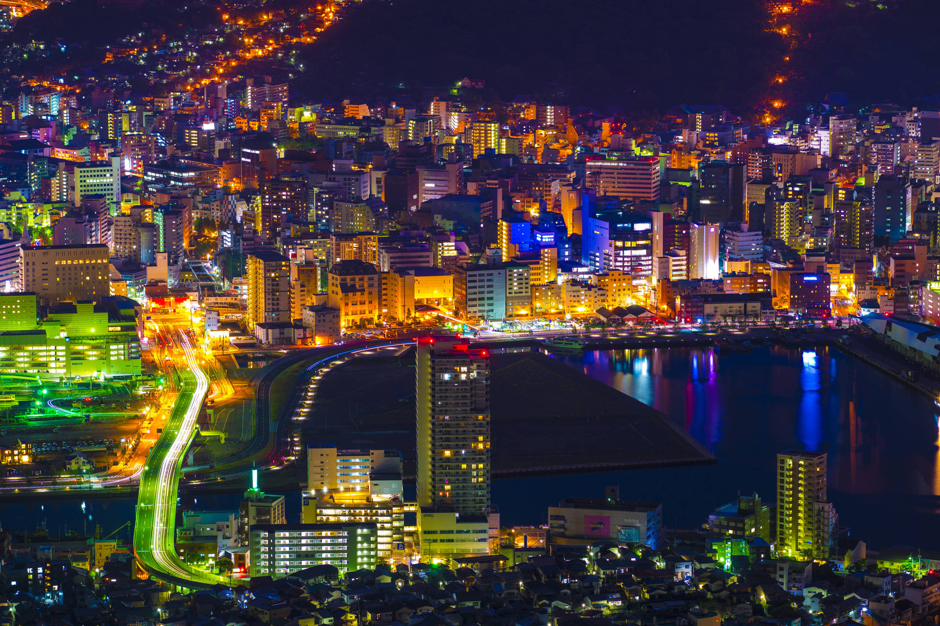 日本絢爛的夜空 壁紙 — 一個耀眼的夜間城市景觀，閃爍的霓虹燈光倒映在水面上，車流的光跡照亮蜿蜒的道路。