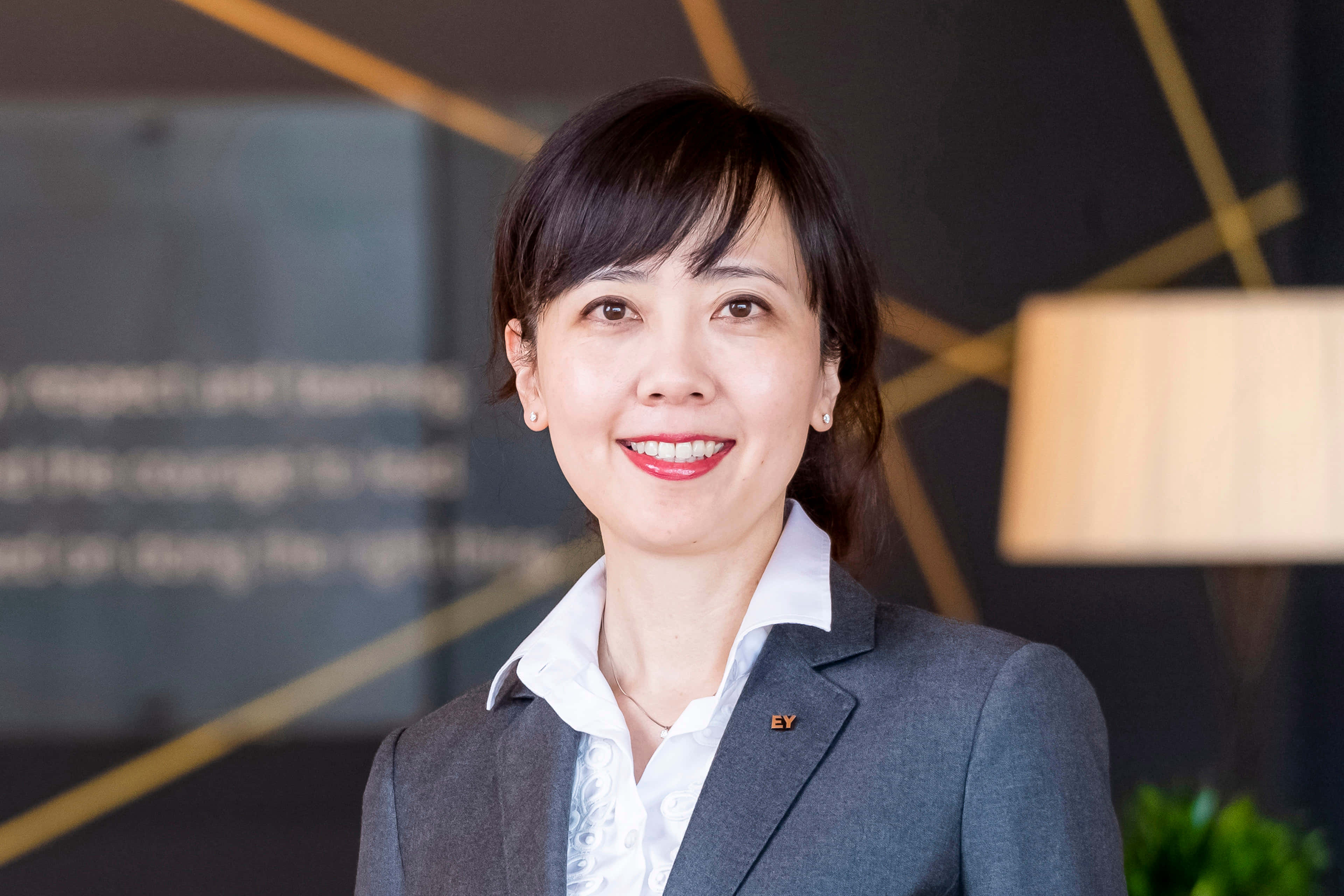 日本綜合格鬥選手 Megumi Fujii 專業造型 壁紙 — 一位微笑的女商人穿著灰色西裝和白色襯衫，自信地站在一個現代辦公室，帶有幾何金色牆壁裝飾。
