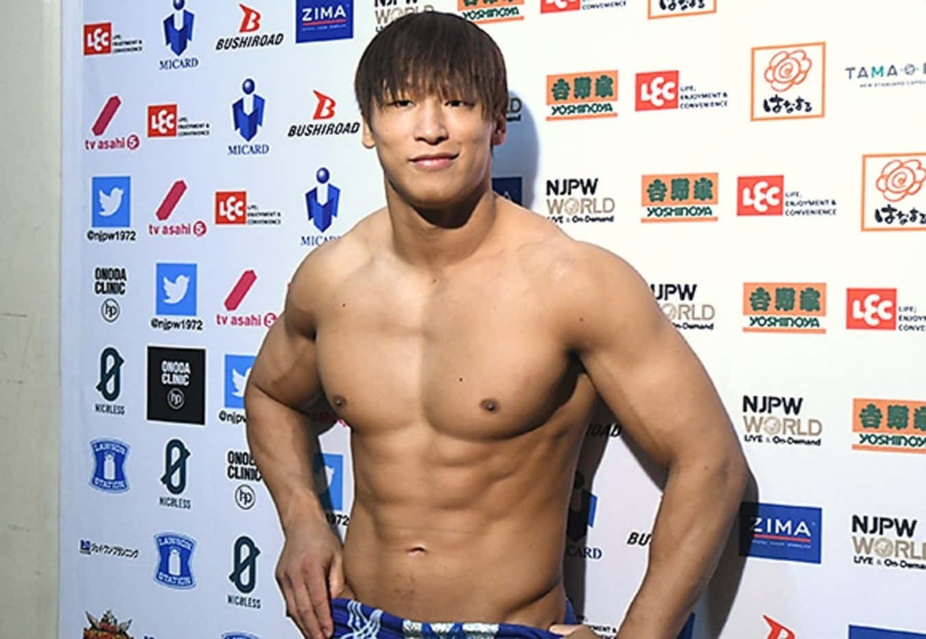 日本職業摔角手 Kota Ibushi 舉行記者會 壁紙 — 一個肌肉發達、冇著衫嘅短黑髮男人自信地企喺一個有標誌同日文嘅品牌活動背景前面。