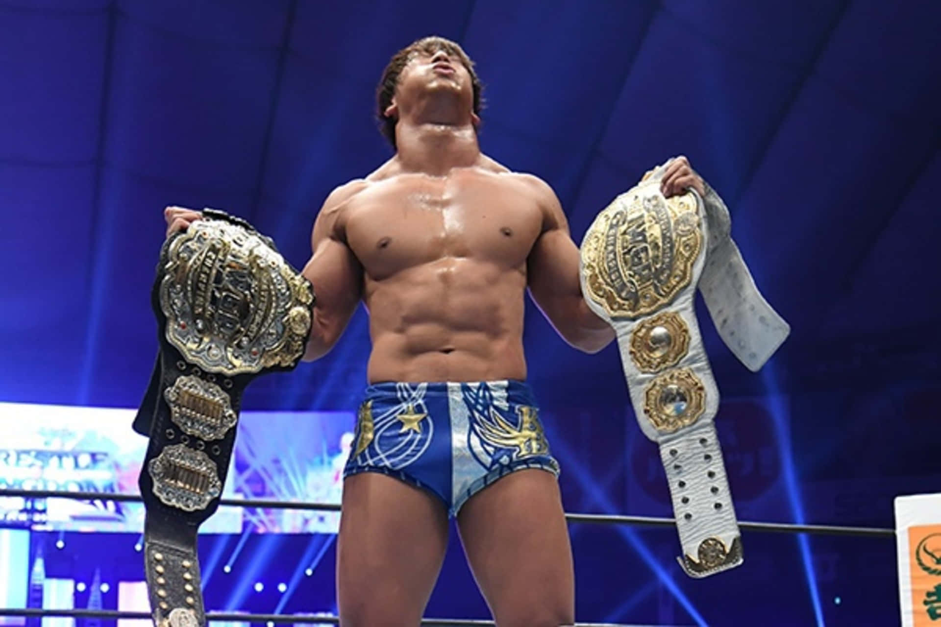 日本職業摔角手 Kota Ibushi，2021 年 IWGP 國際冠軍 壁紙 — 一位肌肉發達的摔角手在明亮的場館燈光下，勝利地站在擂台上高舉兩條冠軍腰帶。