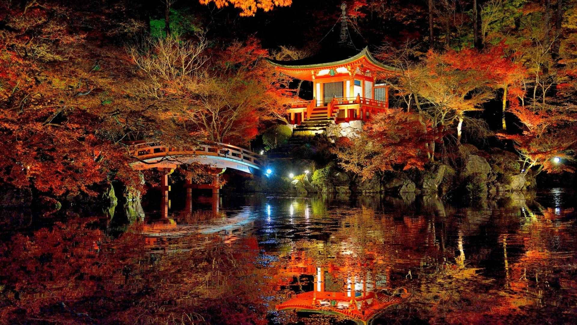 日本自然中的寺廟 壁紙 — 夜晚，一座發光的紅色日本寺廟和拱橋倒映在平靜的水面上，上方是火紅的秋樹。