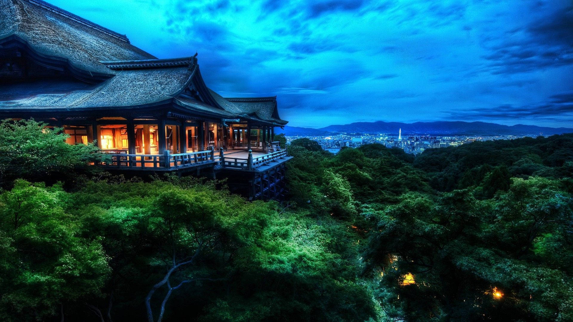 日本自然中的寺廟森林 壁紙 — 一座傳統日本寺廟坐落在茂密綠樹之上，俯瞰著深藍色暮色天空下的廣闊城市景觀。