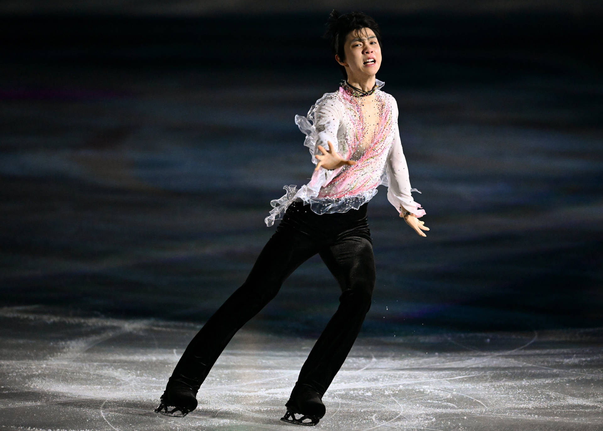 日本花式滑冰選手羽生結弦於冬季奧運會 壁紙 — 一位身穿閃亮白色和粉紅色服裝的花樣滑冰選手，在戲劇性的舞台燈光下，動態地滑過冰場。