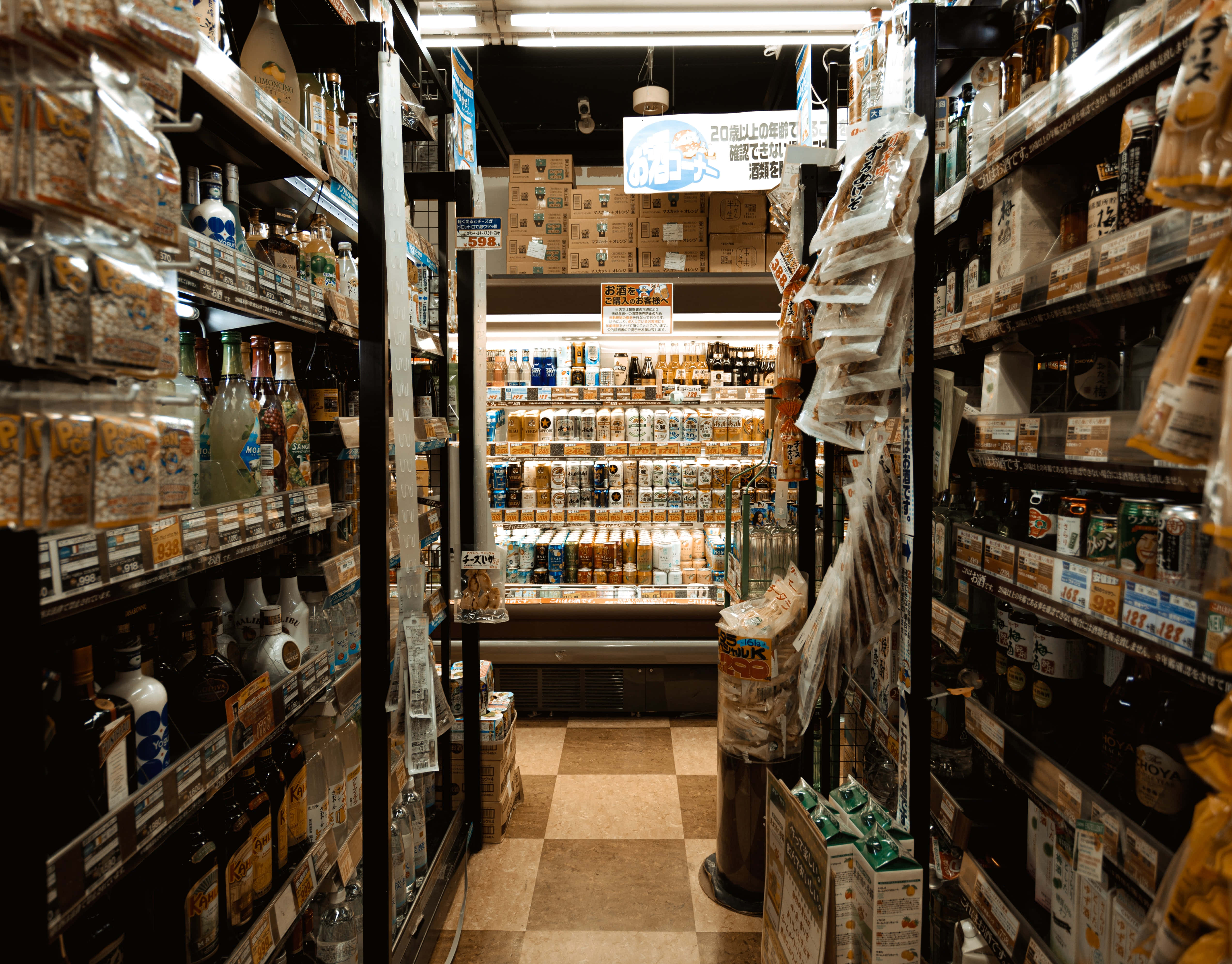 Grocery Store 桌布 壁紙 — 日本酒舖內一條狹窄的通道，兩旁擺滿了瓶子和包裝商品的貨架，上方是明亮的螢光燈。