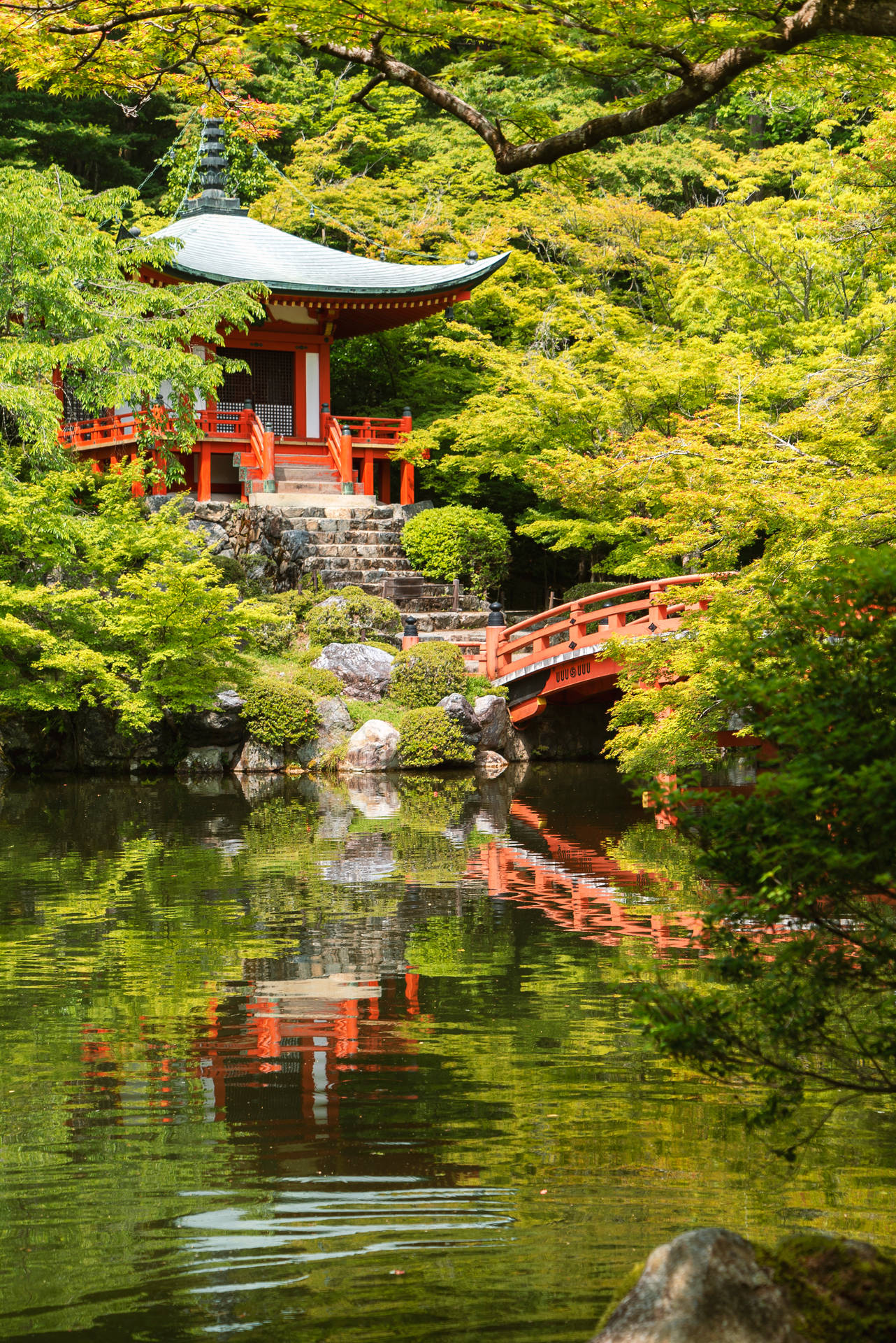 紅色寺廟和橋樑倒映在寧靜的池塘中，周圍環繞著綠樹。