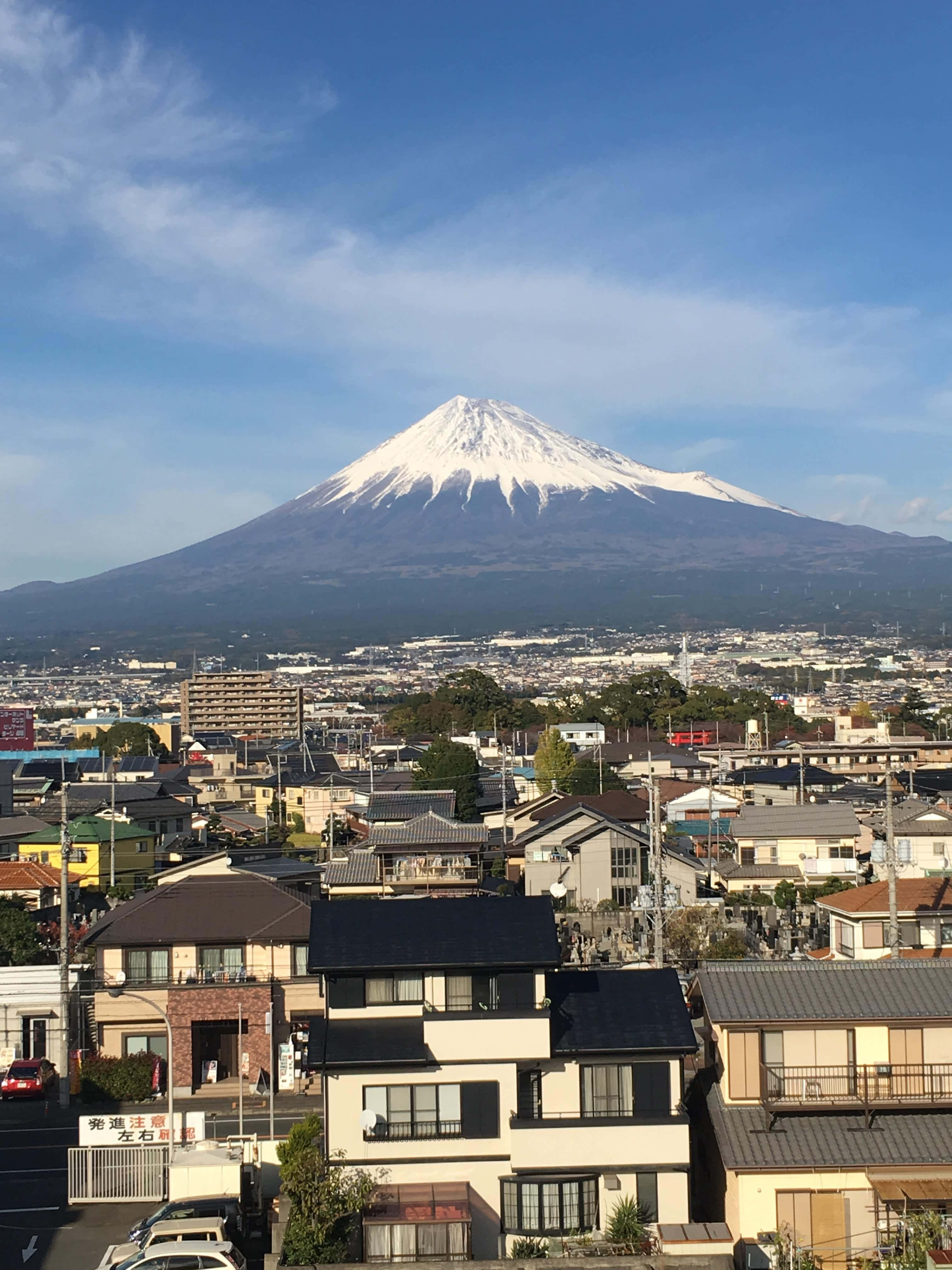 日本雪頂富士山的驚豔之美 壁紙 — 白雪皚皚的富士山聳立在一個廣闊的日本城鎮之上，天空湛藍清澈，飄著縷縷薄雲。