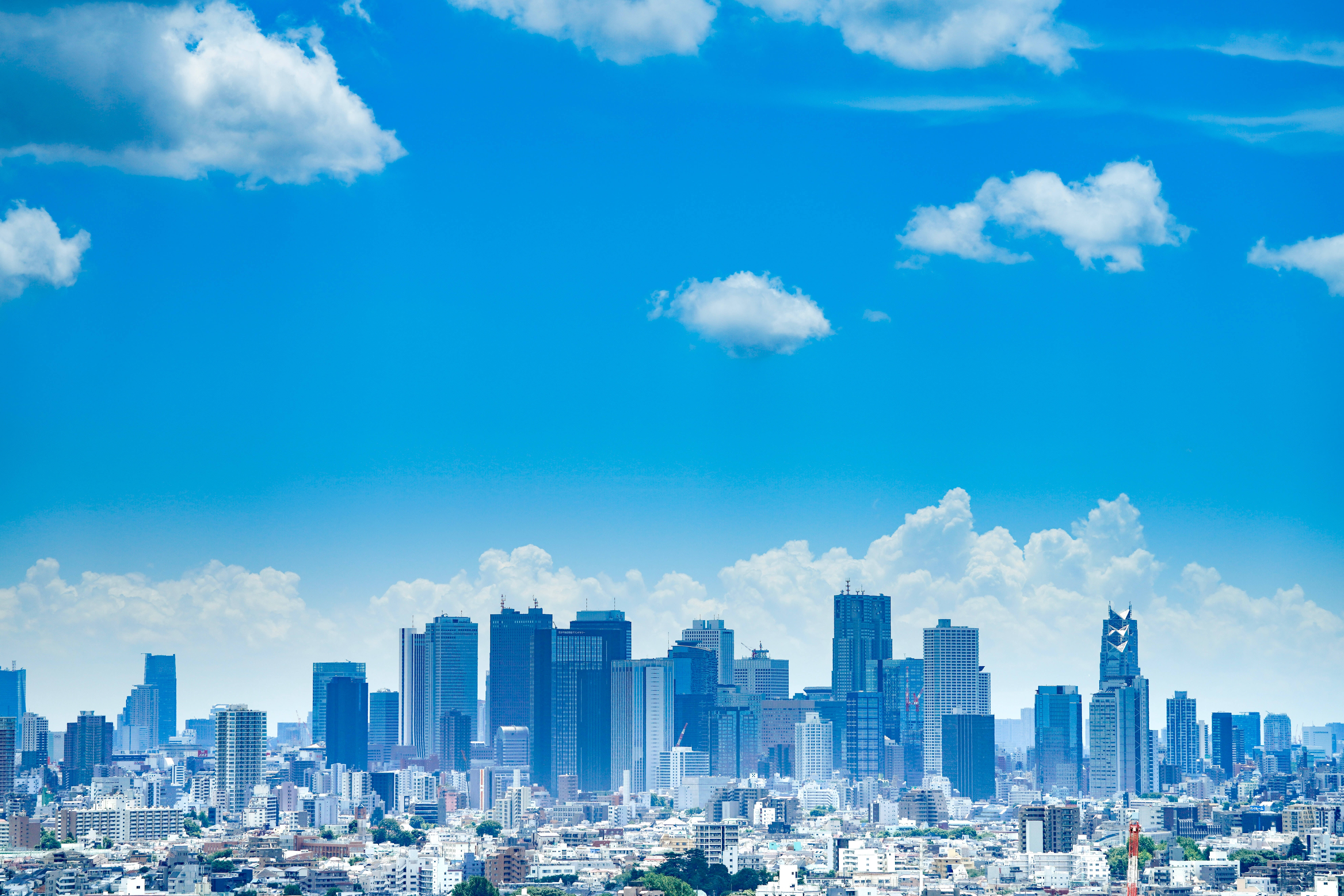 日本高畫質東京天際線 壁紙 — 明亮的藍天與蓬鬆的白雲籠罩著密集的現代城市景觀，包括玻璃摩天大樓和低層建築。