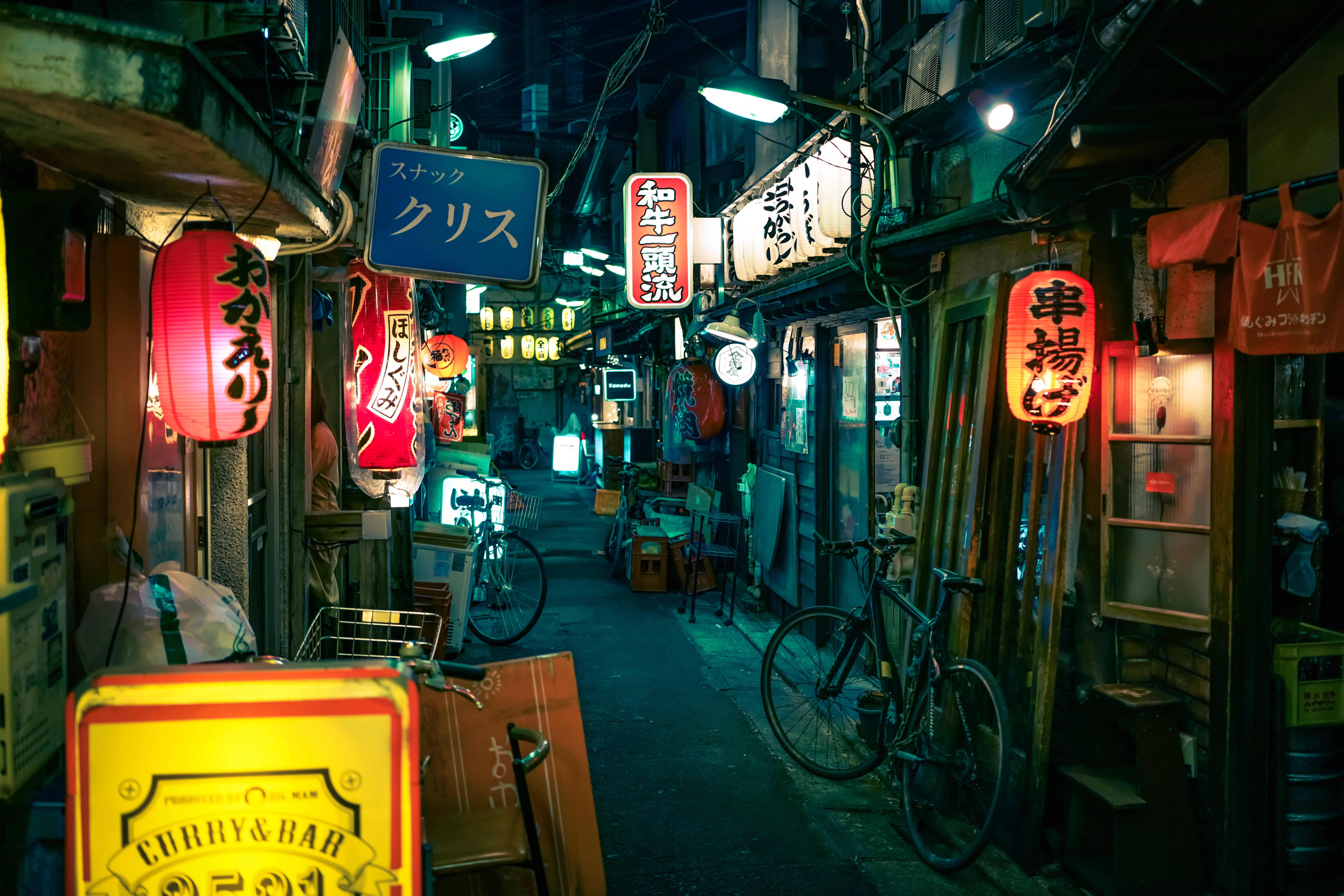日本高畫質東京巷弄餐廳 壁紙 — 日本夜晚一條狹窄的霓虹燈小巷，有發光的招牌、單車和木製店面，在潮濕的路面上投射出色彩繽紛的倒影。