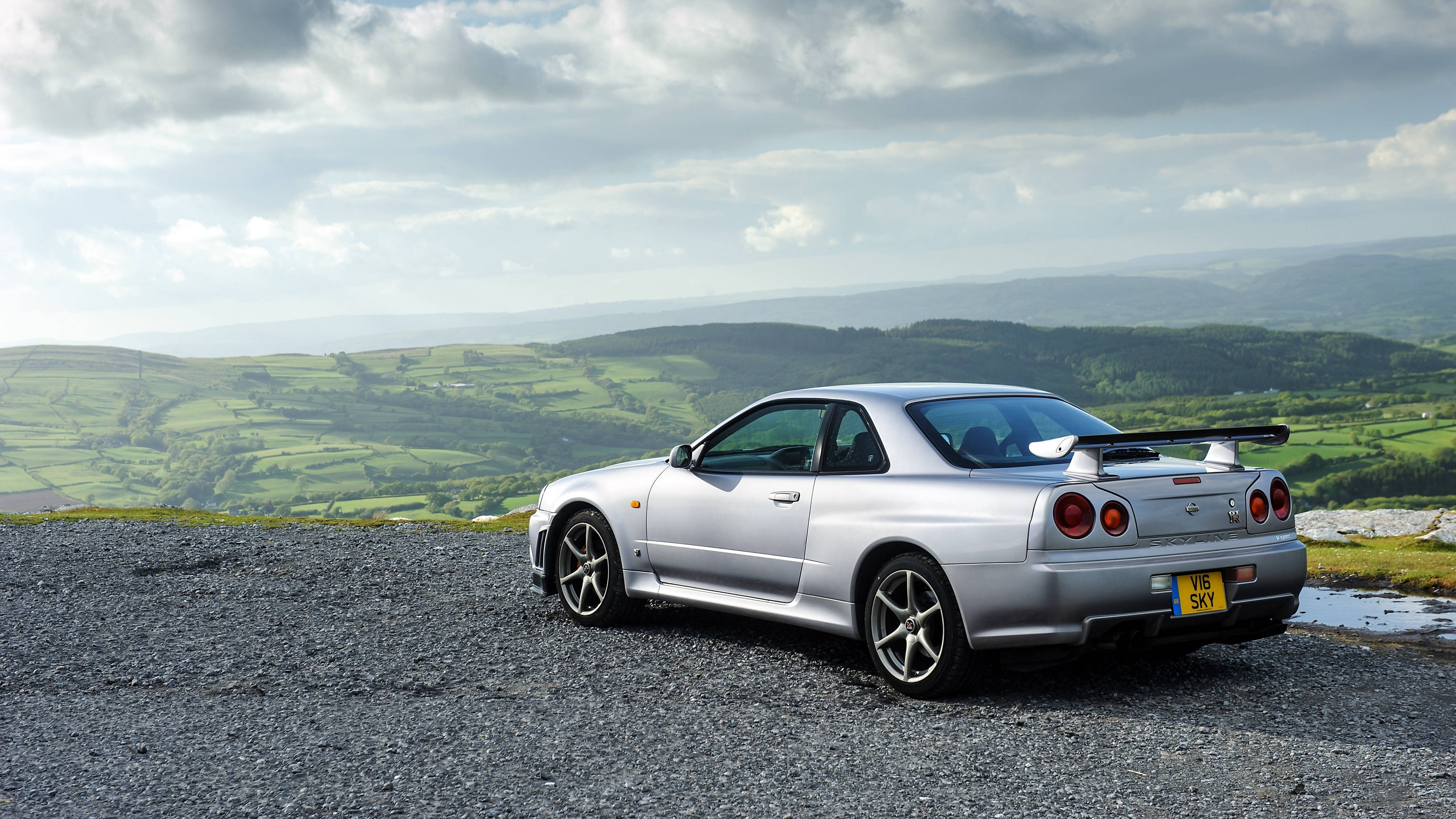 日產GT-R銀色Skyline 壁紙 — 一輛銀色日產Skyline GT-R停泊在碎石觀景點，背景是連綿起伏的綠色山丘和多雲的天空。