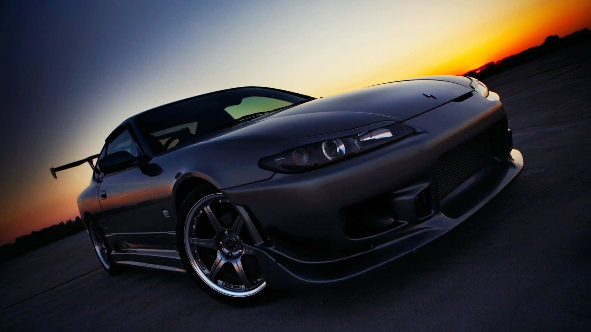 日產Silvia S15 天空視角 壁紙 — 一輛時尚的啞光黑色跑車帶有後翼，在日落時分傾斜在開闊的道路上，天空呈現溫暖的橙色和涼爽的藍色。