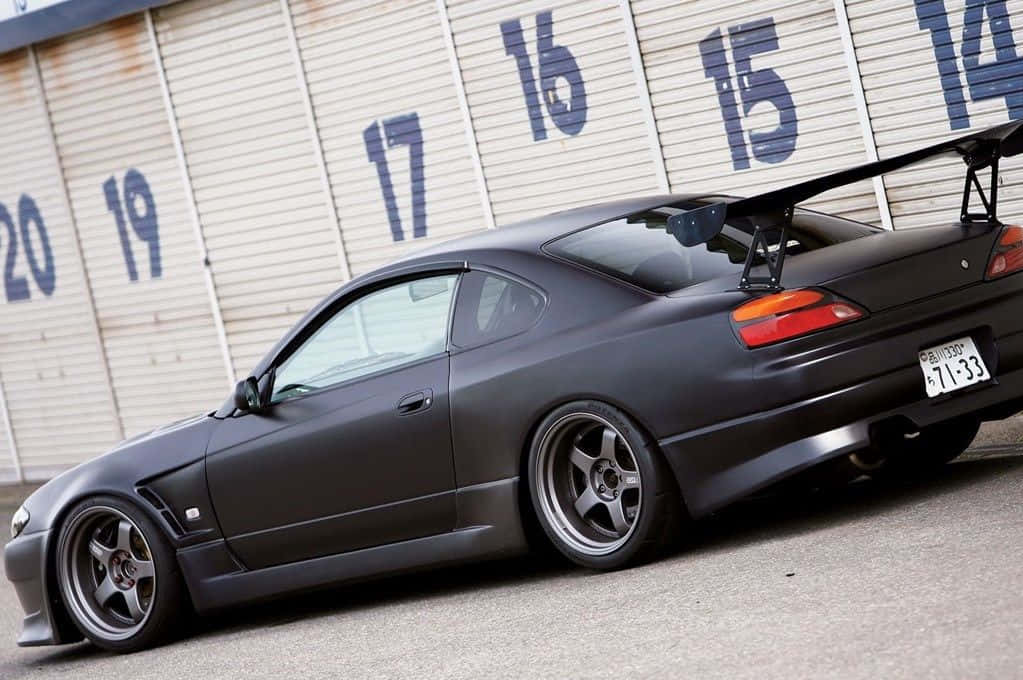 日產Silvia S15超黑版 壁紙 — 一架啞黑色跑車帶有大型尾翼，停泊在帶有編號的倉庫門前。