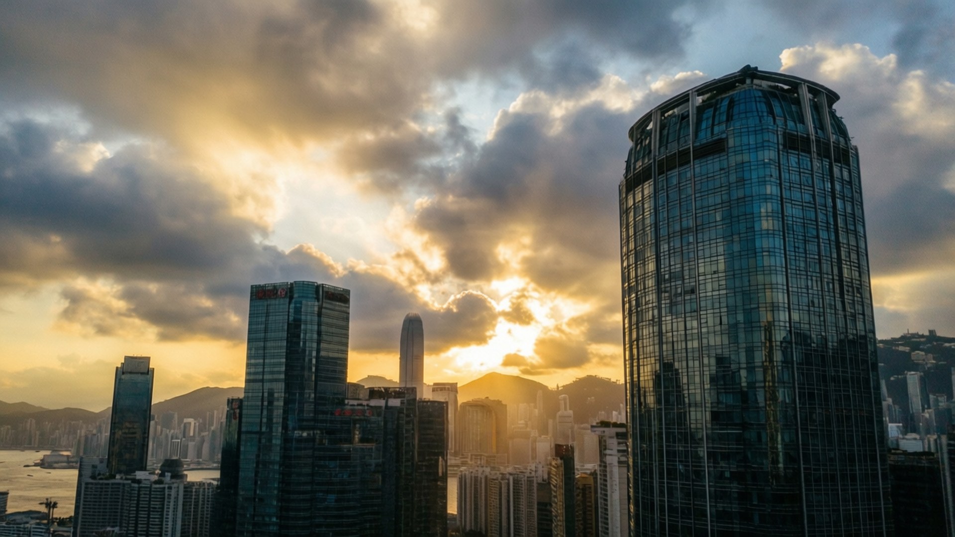 日落塔樓全景 壁紙 — 夕陽穿過戲劇性的雲層，照耀在一群密集的玻璃摩天大樓上，反射出金色的光芒。