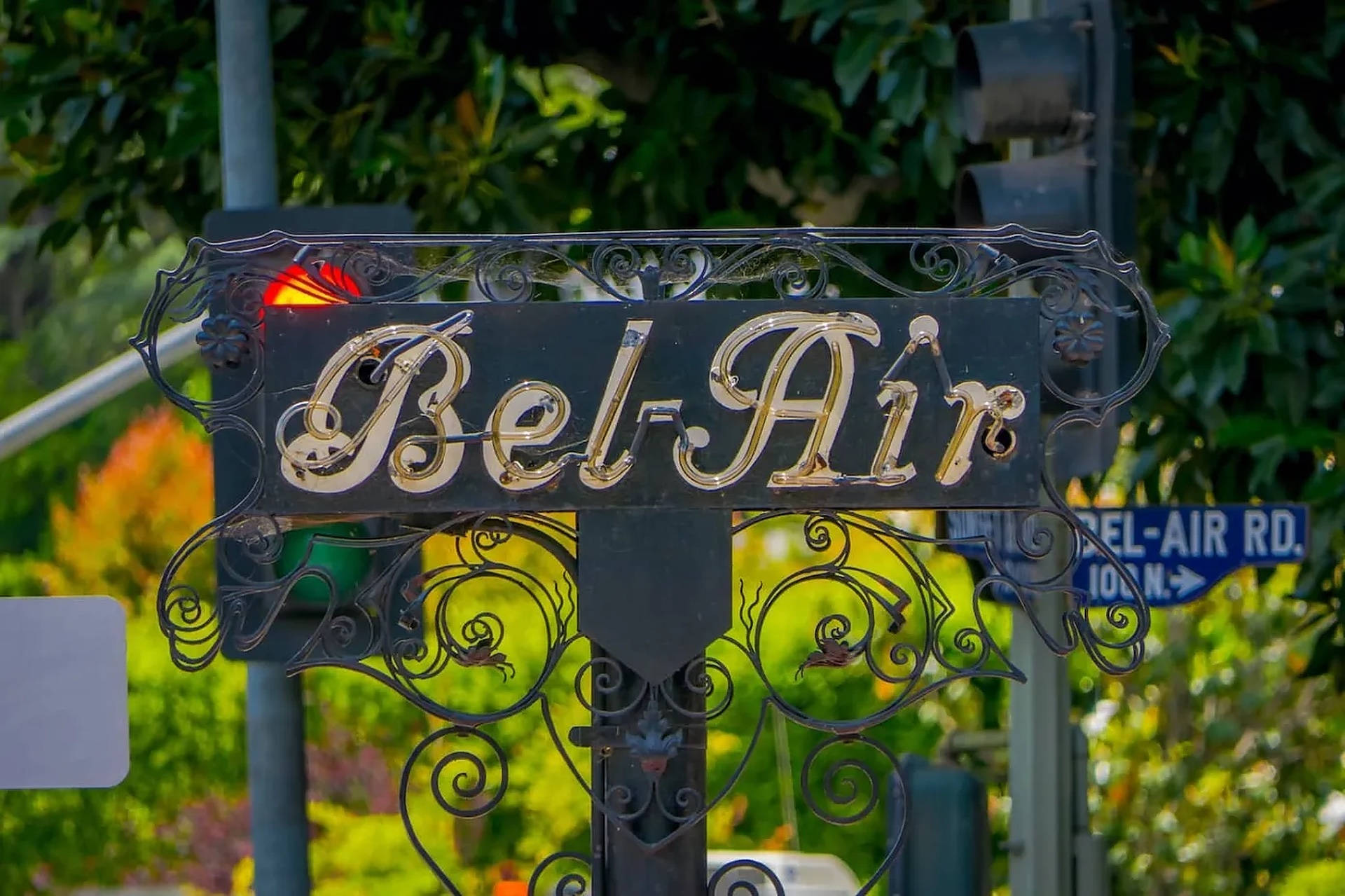 日落大道上的貝萊爾 壁紙 — 一個裝飾性的黑色鍛鐵標誌，帶有金色 'Bel-Air' 字樣，矗立在街角，周圍環繞著茂密的綠色樹葉。