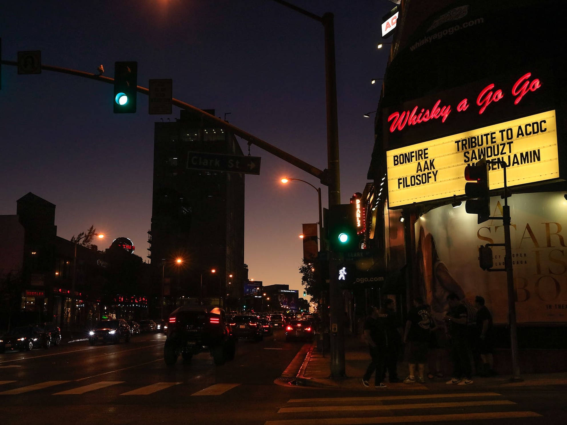 日落大道夜景 壁紙 — 黃昏時分，一條霓虹燈照亮嘅城市街道，發光嘅「Whisky a Go Go」招牌同交通燈喺濕漉漉嘅路面投射出溫暖嘅光芒。