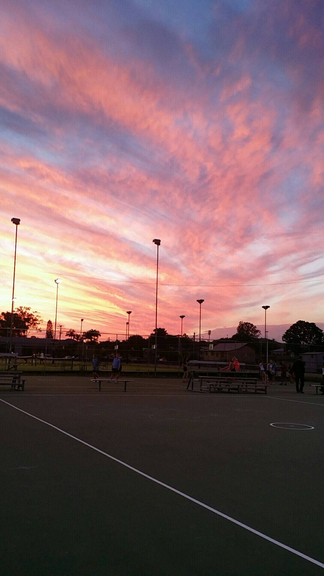 日落大道粉紅天空 壁紙 — 黃昏時分的網球場沐浴在火紅的粉紅色和橙色日落雲層中，帶有剪影的球員和燈光。