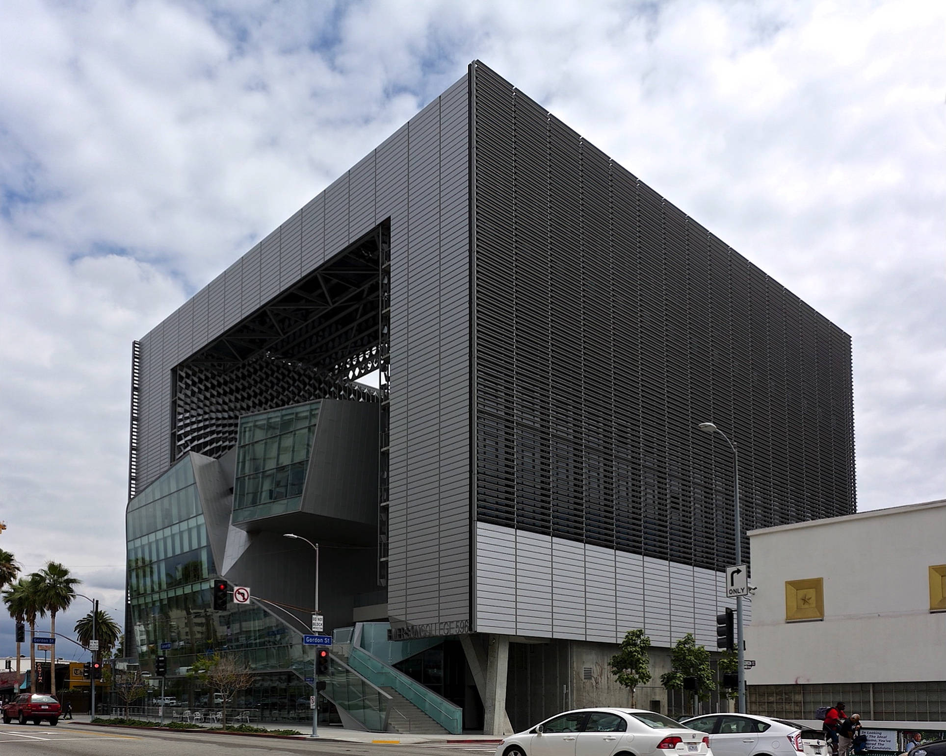 日落大道艾默生學院 壁紙 — 一座現代角形建築，帶有深色金屬板和玻璃部分，矗立在多雲的天空下，附近有汽車和棕櫚樹。