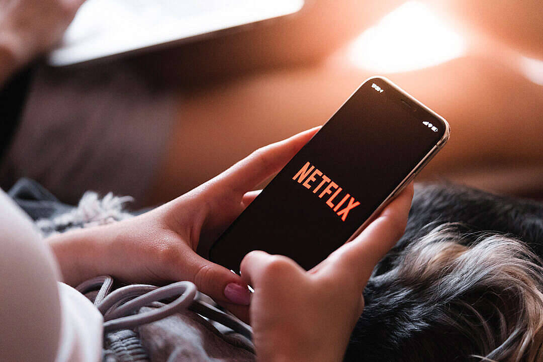 日落山脈風景 壁紙 — 特寫鏡頭：手裡拿著一部顯示Netflix標誌的智能手機，同時在沙發上休息。