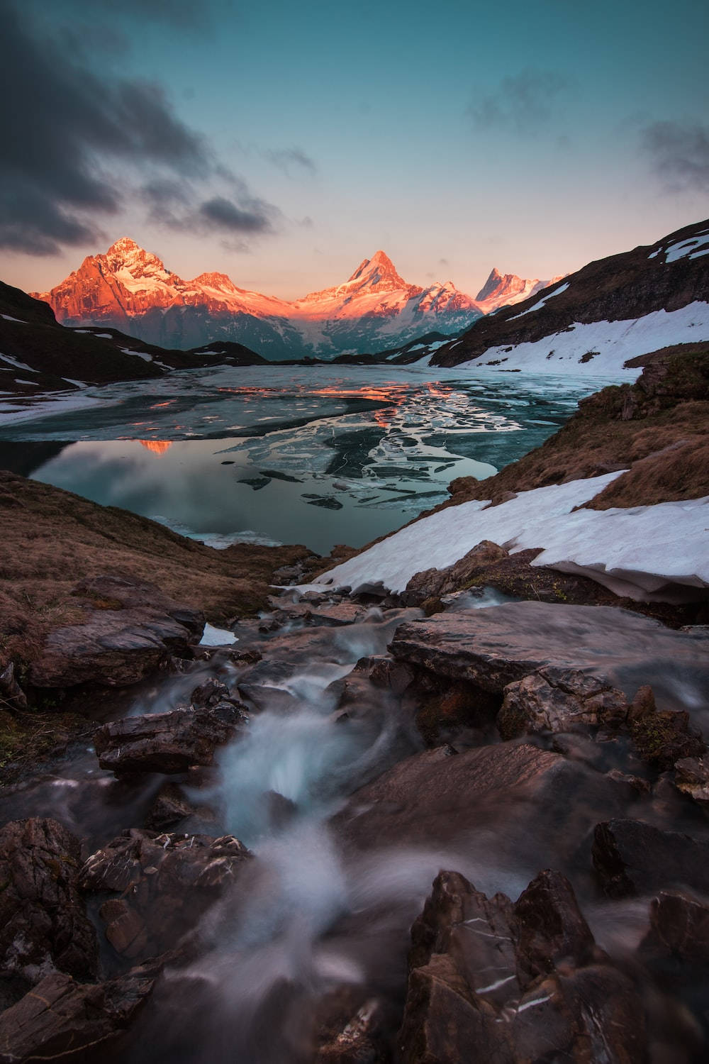 Switzerland 桌布 壁紙 — 日落時分，寧靜的山間湖泊倒映著積雪的山峰，前景中湍急的溪流流過岩石。