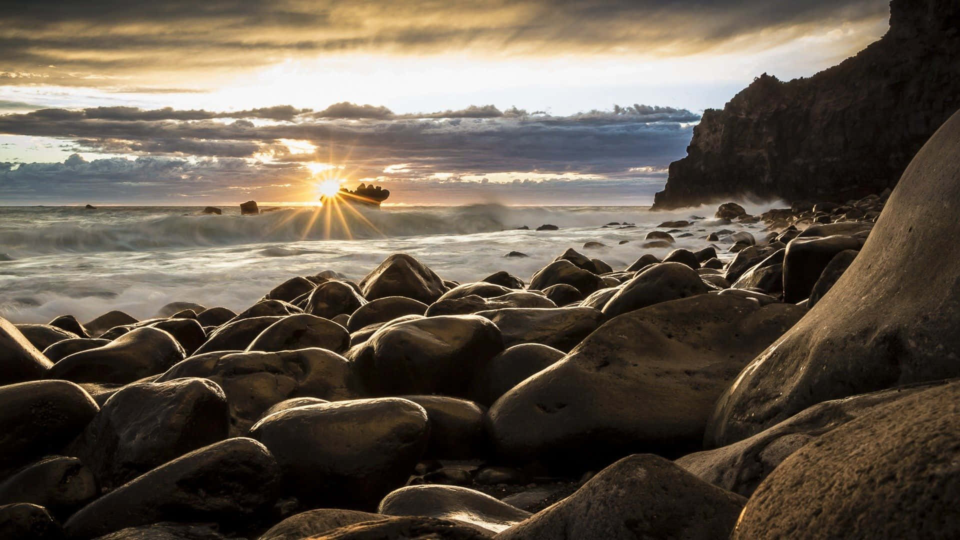 New Zealand 桌布 壁紙 — 日落時分，岩石海岸線，海浪拍打，戲劇性的雲層將金色光芒投射在濕漉漉的石頭上。