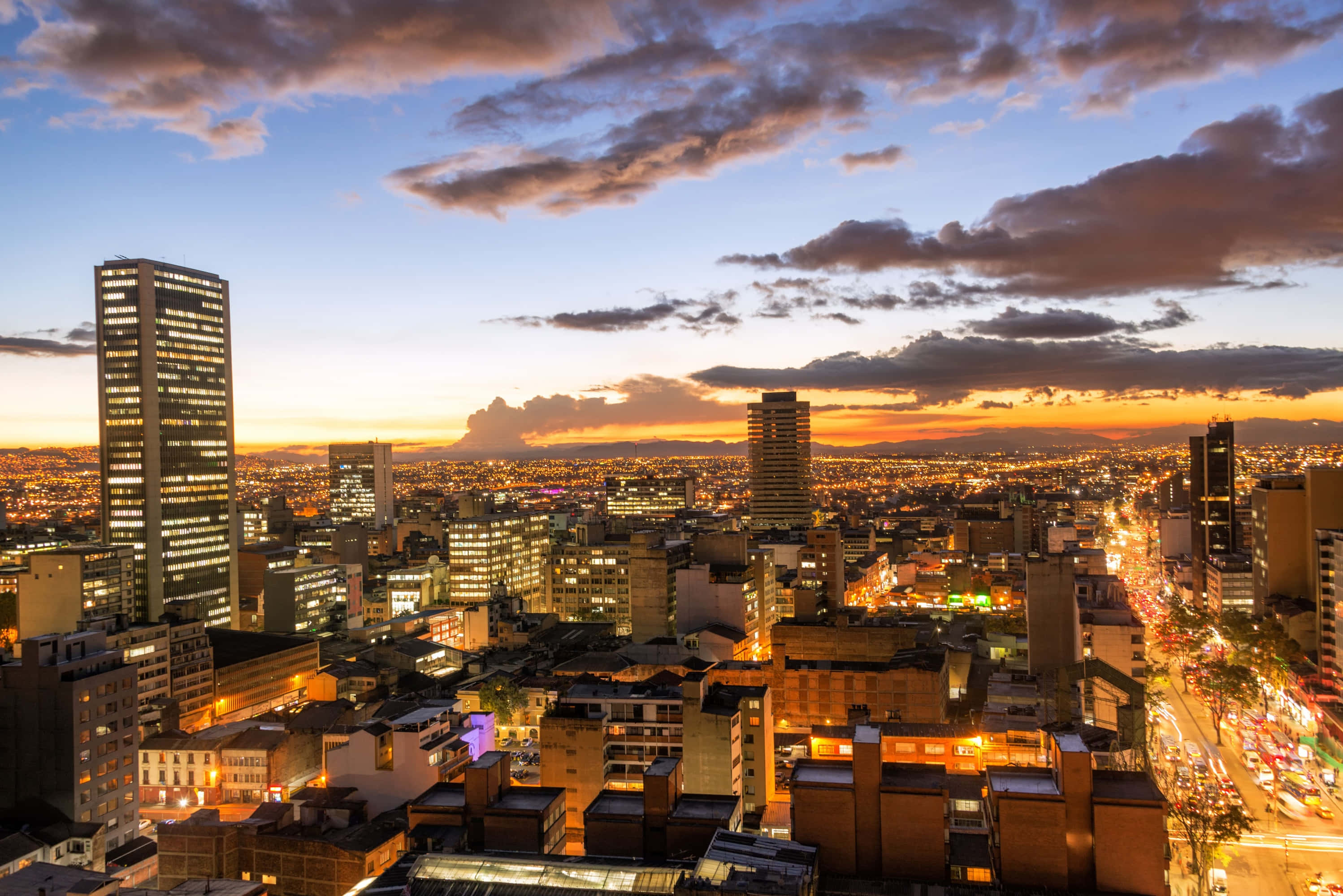Colombia 桌布 壁紙 — 日落時分，廣闊的城市景觀，金色燈光在戲劇性的多雲天空下照亮摩天大樓和街道。