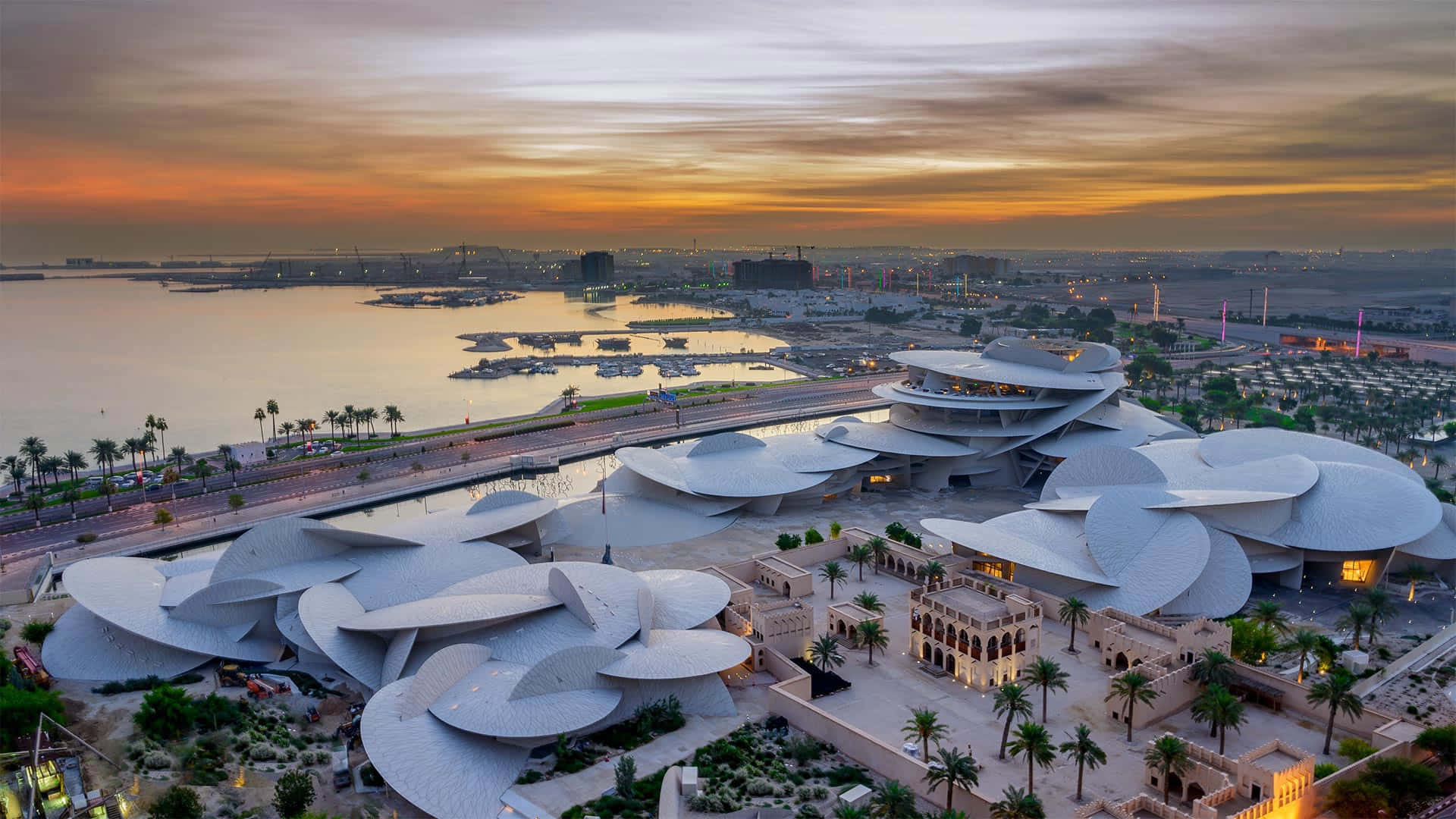 Qatar 桌布 壁紙 — 日落時分，未來主義白色花瓣形建築的鳥瞰圖，旁邊是海濱，有棕櫚樹和遠處的城市燈光。