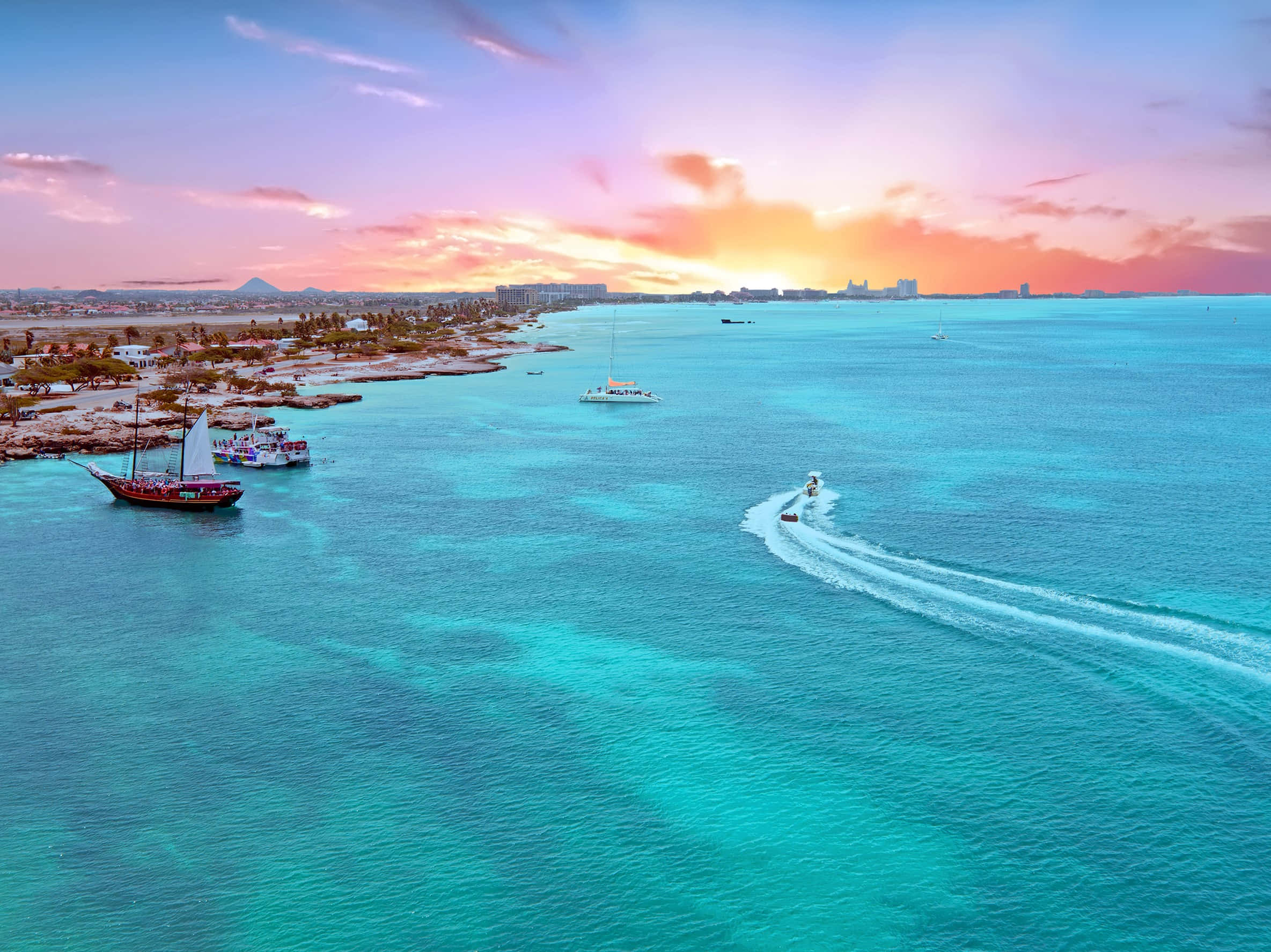 Aruba 桌布 壁紙 — 日落時分，海岸小鎮附近，帆船和快艇留下白色尾跡的碧綠海水鳥瞰圖。