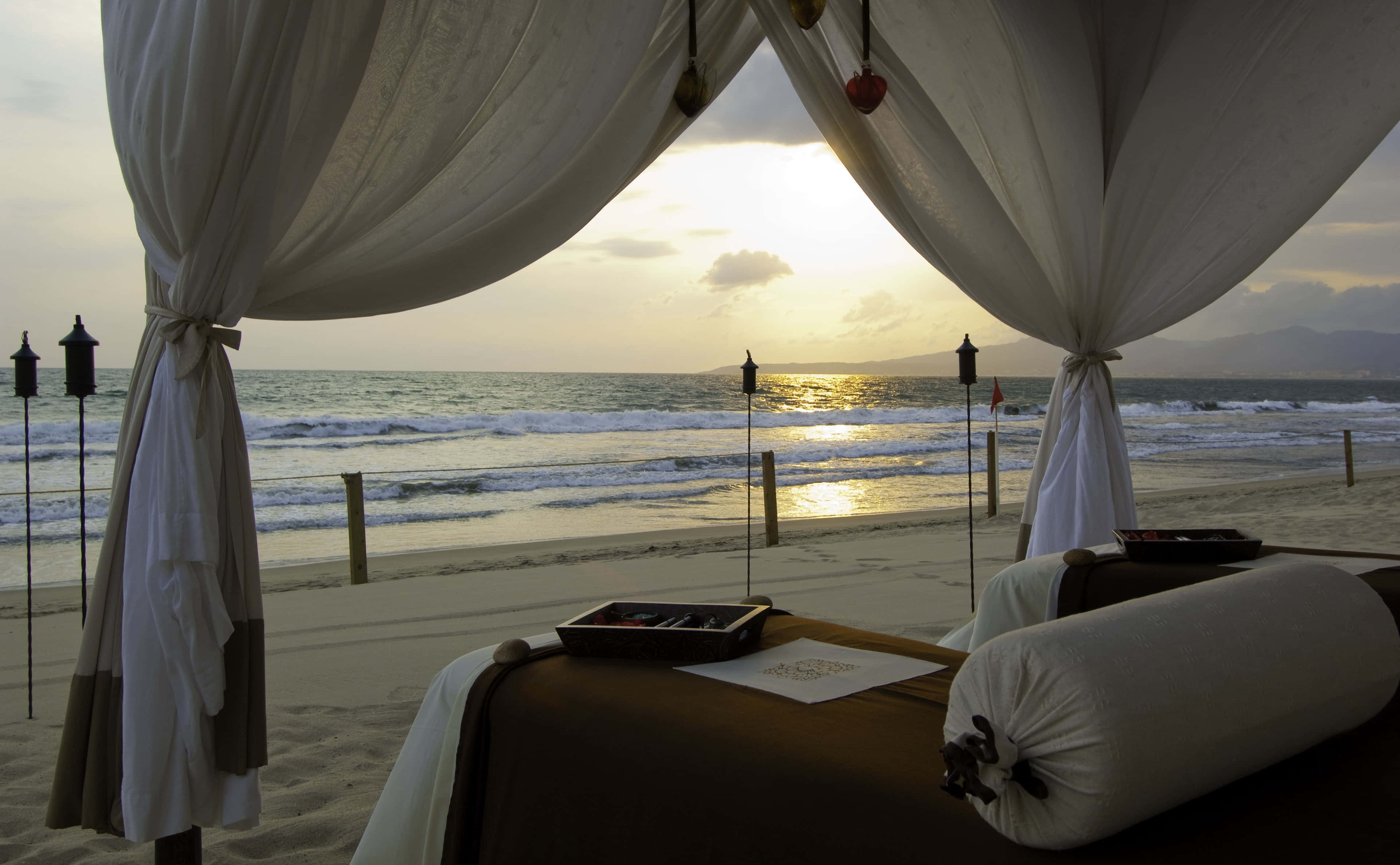 Beach Daybed 桌布 壁紙 — 日落時分，白色天篷下的寧靜海灘按摩床，伴隨著海浪和遠山。