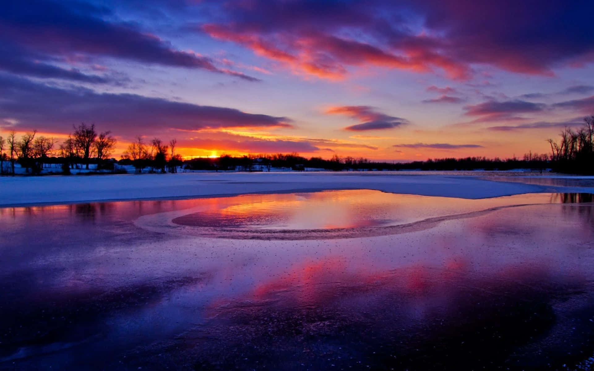 Winter Sunset 桌布 壁紙 — 日落時分，結冰的湖面倒映著火紅橙和紫色的雲彩，映襯著白雪皚皚的冬日景觀。